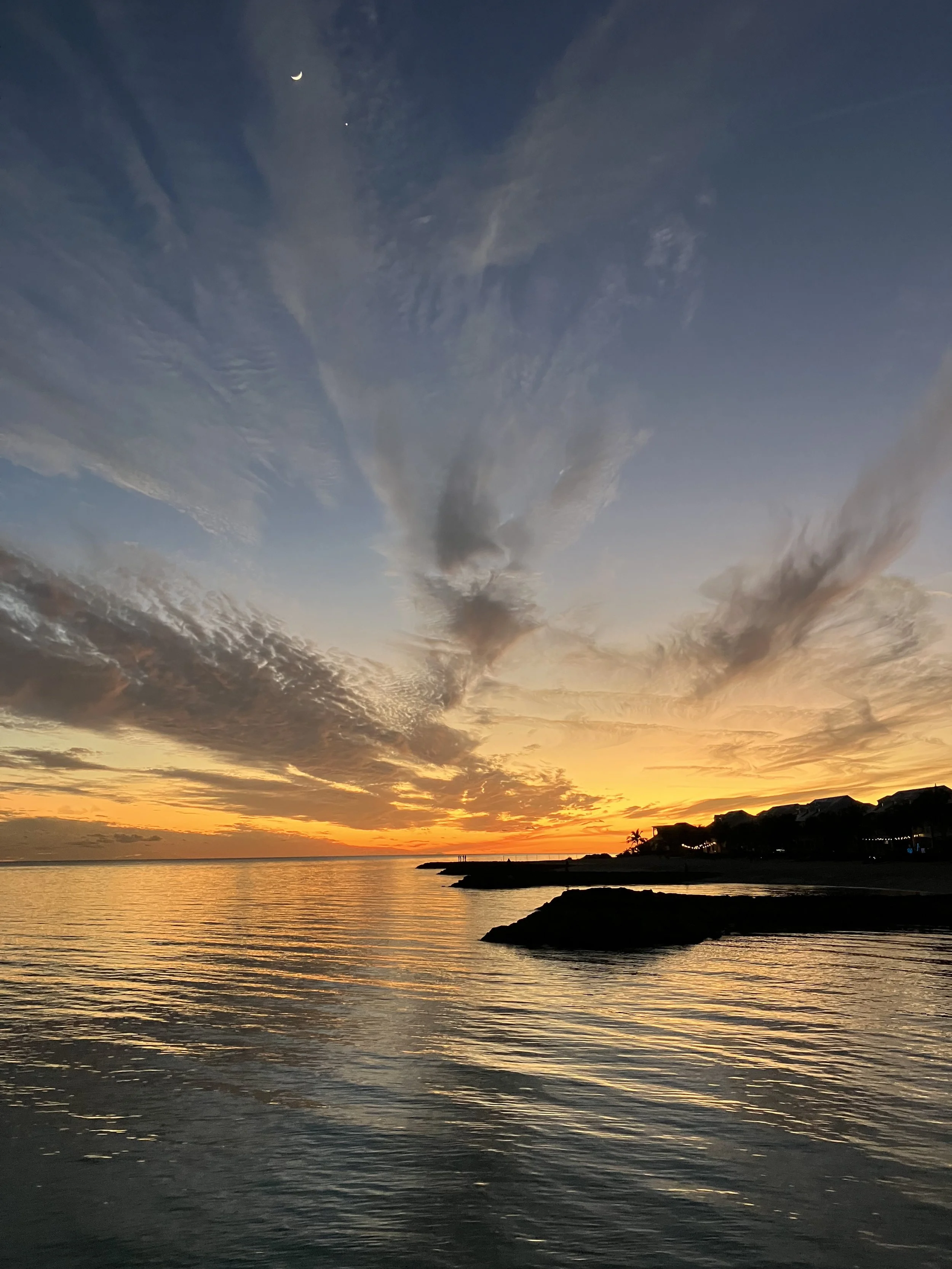 Sunset over the ocean with colorful clouds in the sky and a silhouette of houses and trees on the shoreline.