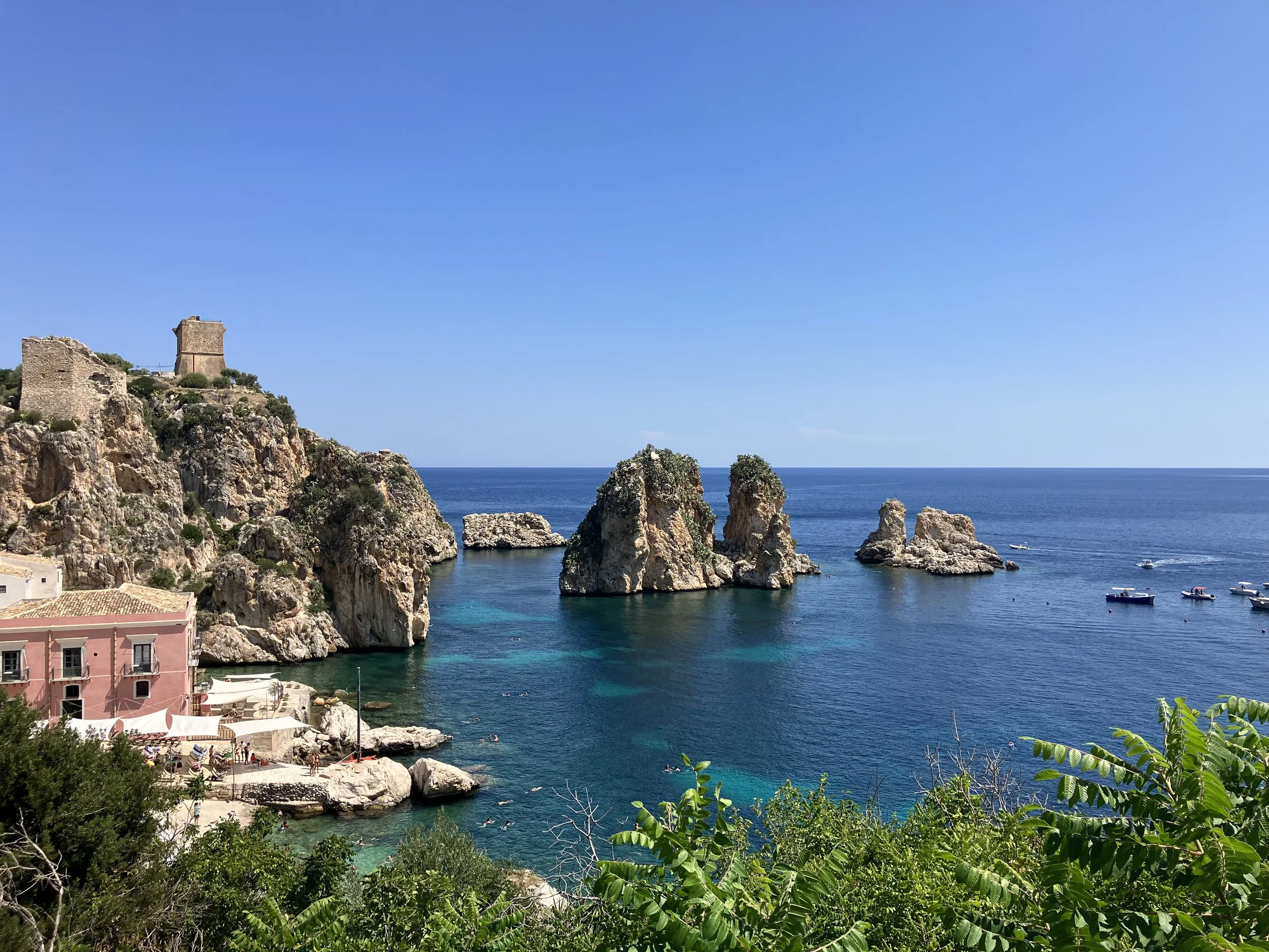 Coastal view of Scopello, Italy with a historic tower and pink building, green foliage in foreground, blue sea with boats, and clear sky in the background.