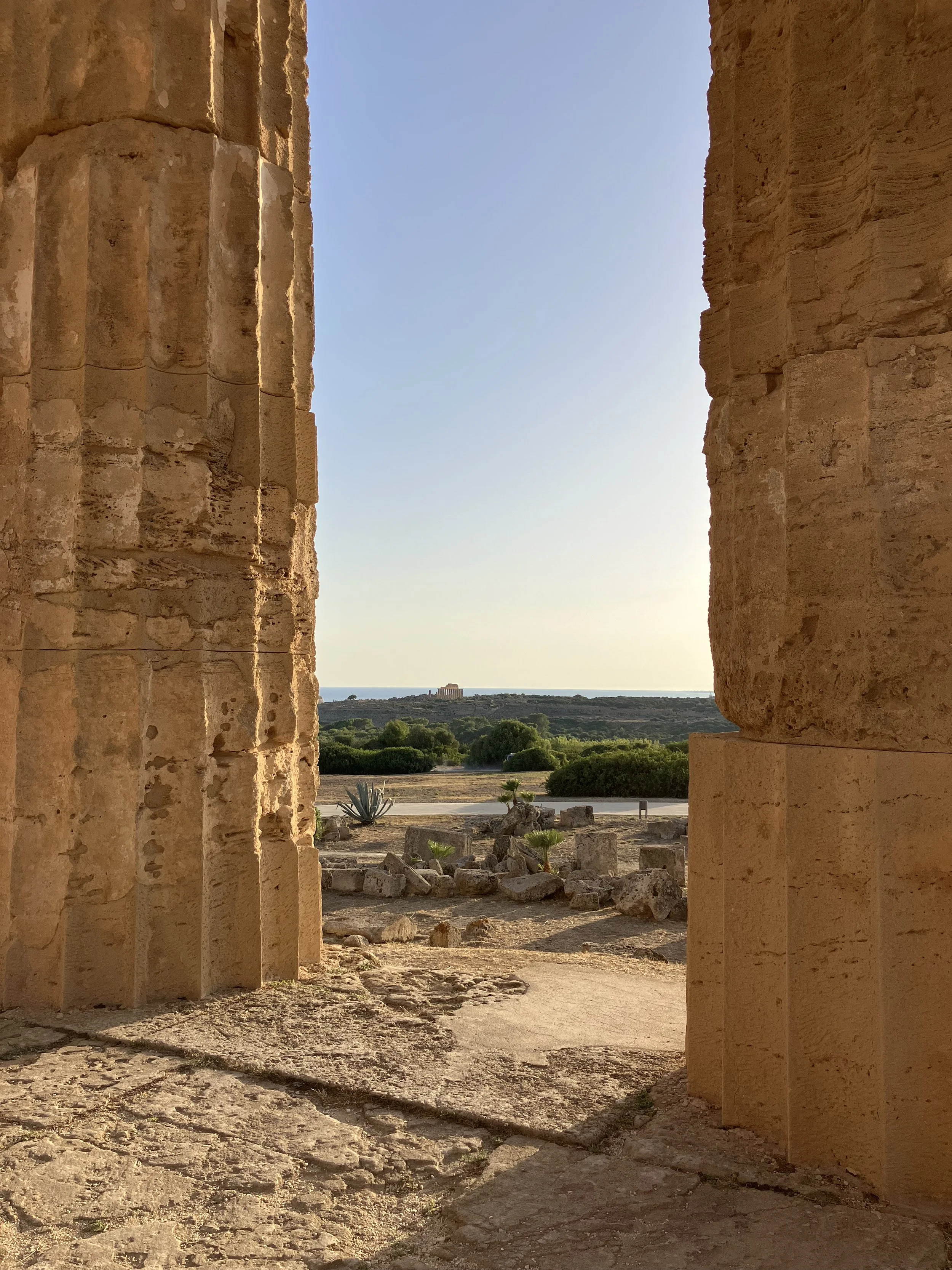 Ancient stone ruins framed by two columns with a view of a landscape featuring greenery, a distant building, and the sea in the background.
