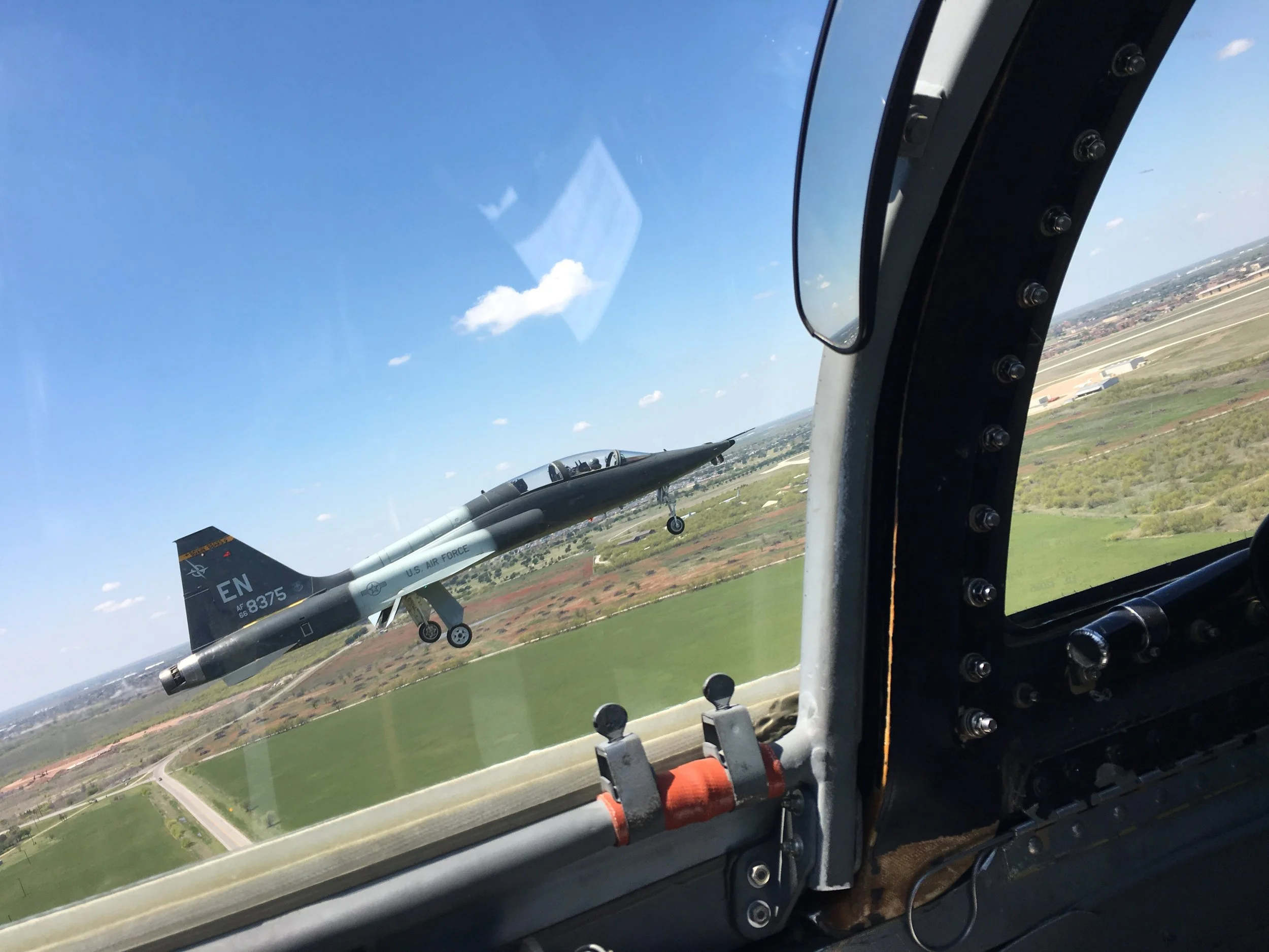 Inside a cockpit of a fighter jet with a view of another fighter jet flying nearby against a blue sky with some clouds and a landscape below.