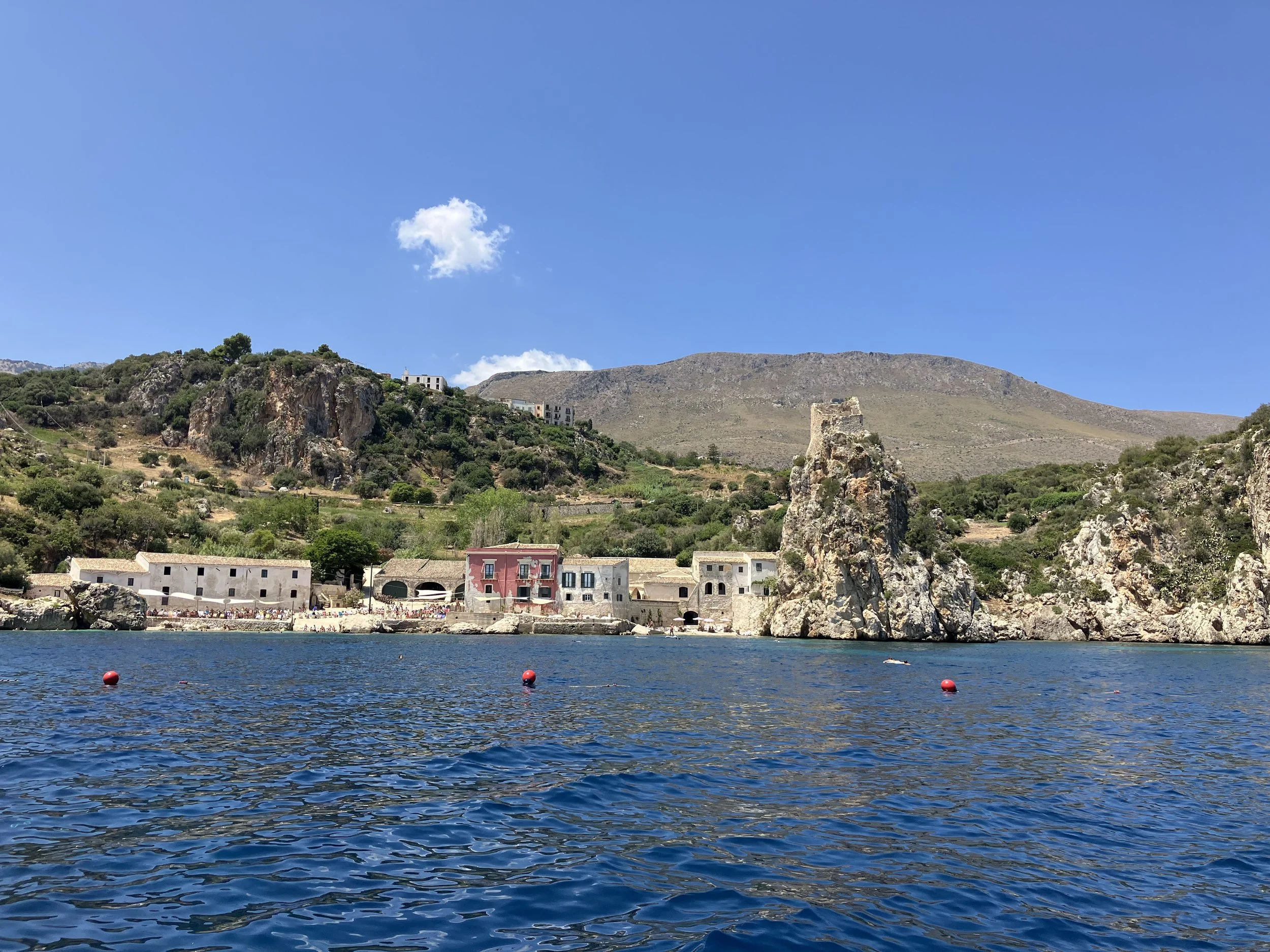 A coastal landscape featuring rocky cliffs, a hillside with scattered buildings, and a body of water in the foreground with red buoys, under a clear blue sky with a few white clouds.