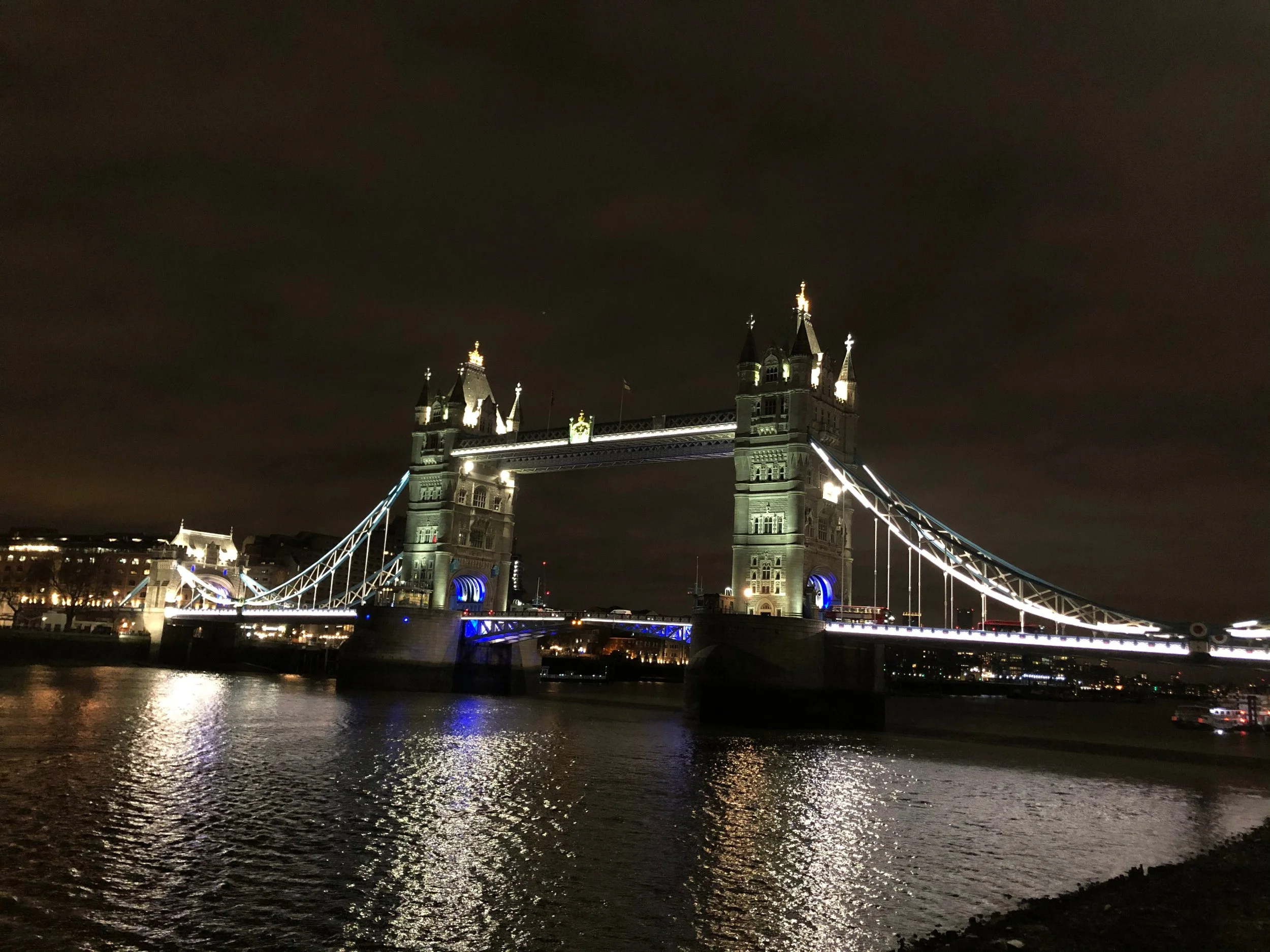 Night view of Tower Bridge in London illuminated with lights, reflecting on River Thames.