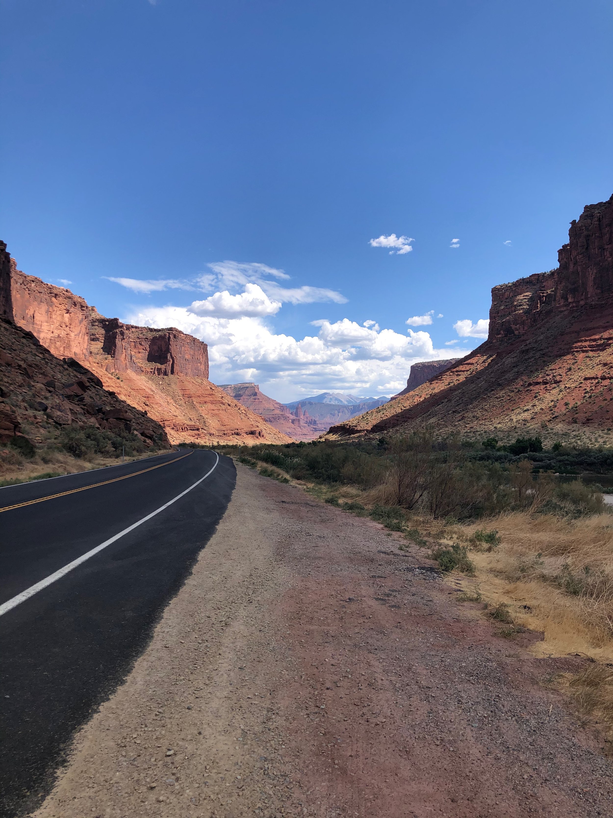 A clear two-lane road running through a desert canyon landscape with reddish rock formations on both sides and a blue sky with scattered white clouds above.