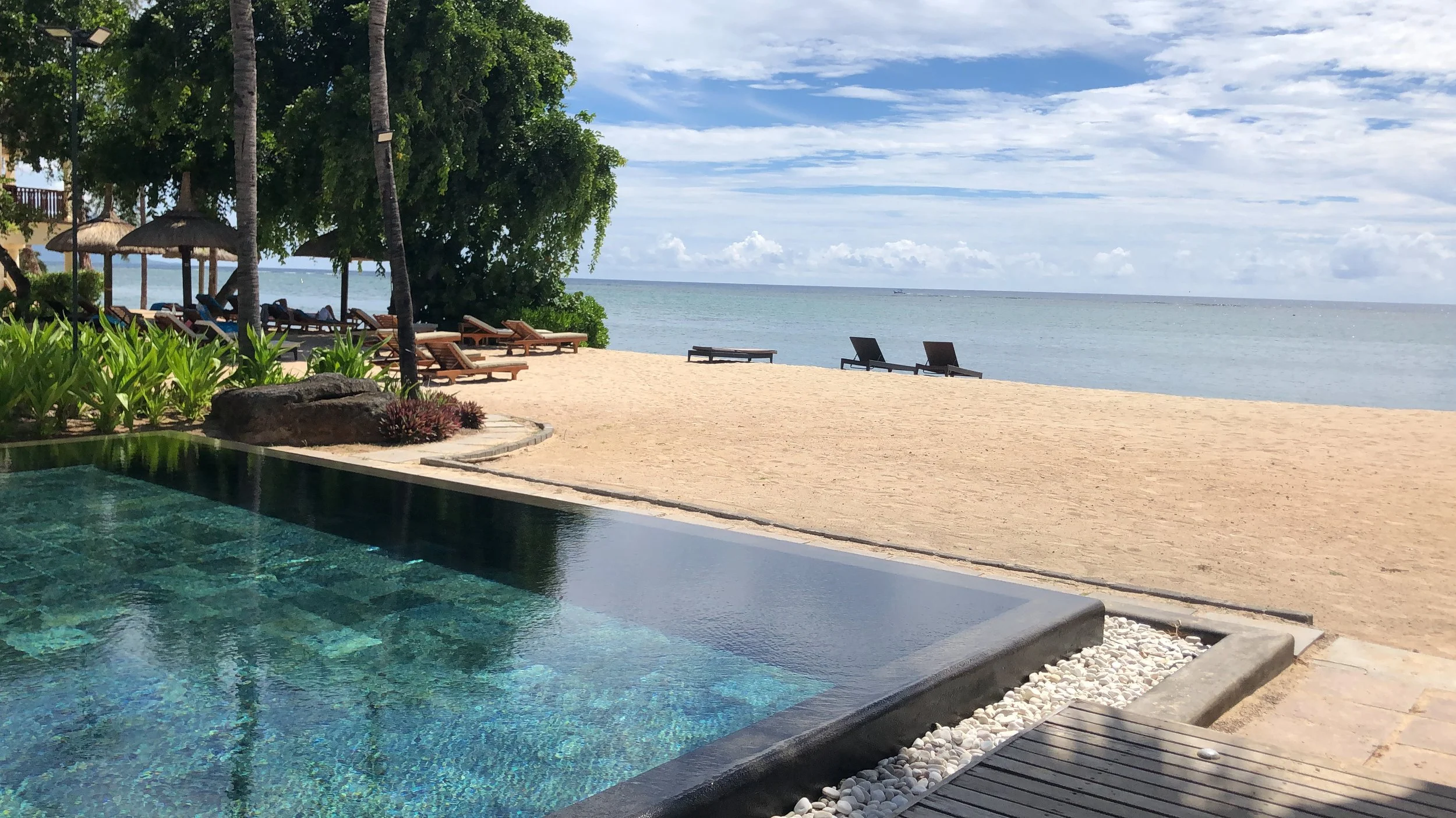 Beachside scene in Mauritius with a swimming pool in the foreground, sandy beach with lounge chairs and umbrellas, calm ocean water, and a partly cloudy sky.