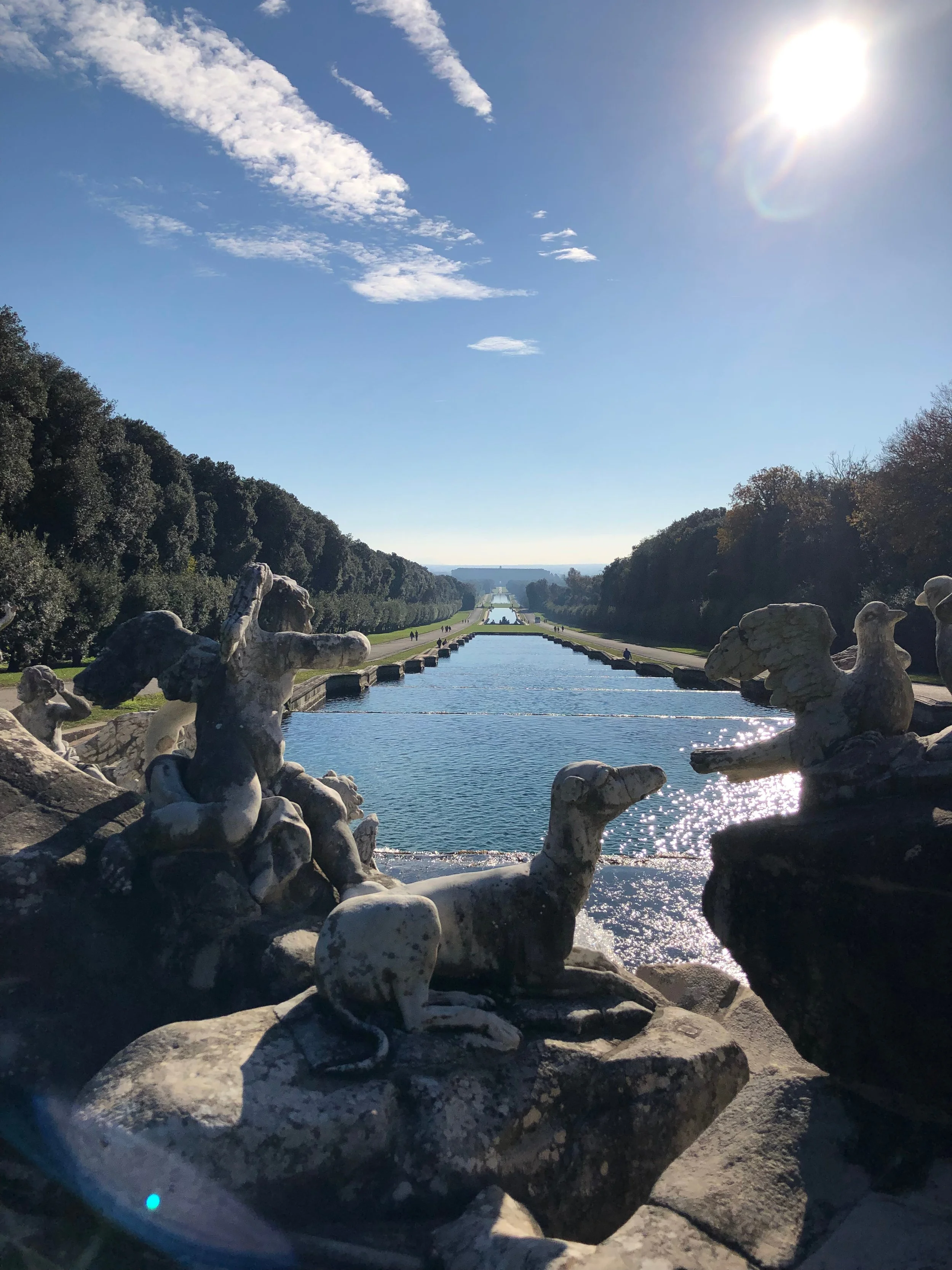The image features a long reflecting pool or canal stretching into the distance, flanked by trees on each side. In the foreground, there are decorative stone sculptures and rocks. The sky is clear with a few clouds, and the sun is shining brightly, c