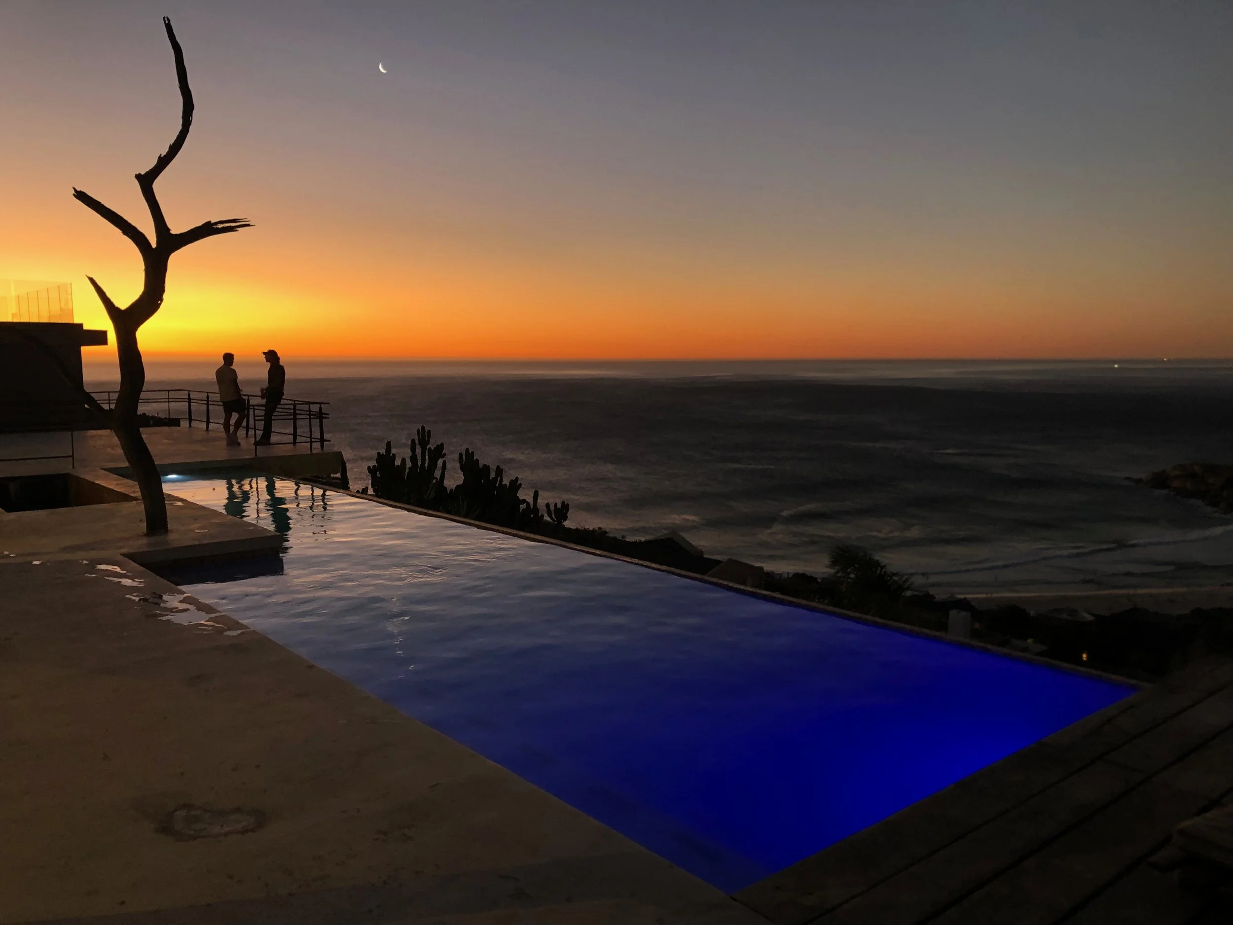 View of a sunset over the ocean with a crescent moon in the sky, a leafless tree in the foreground, and two people standing by a railing near a pool.