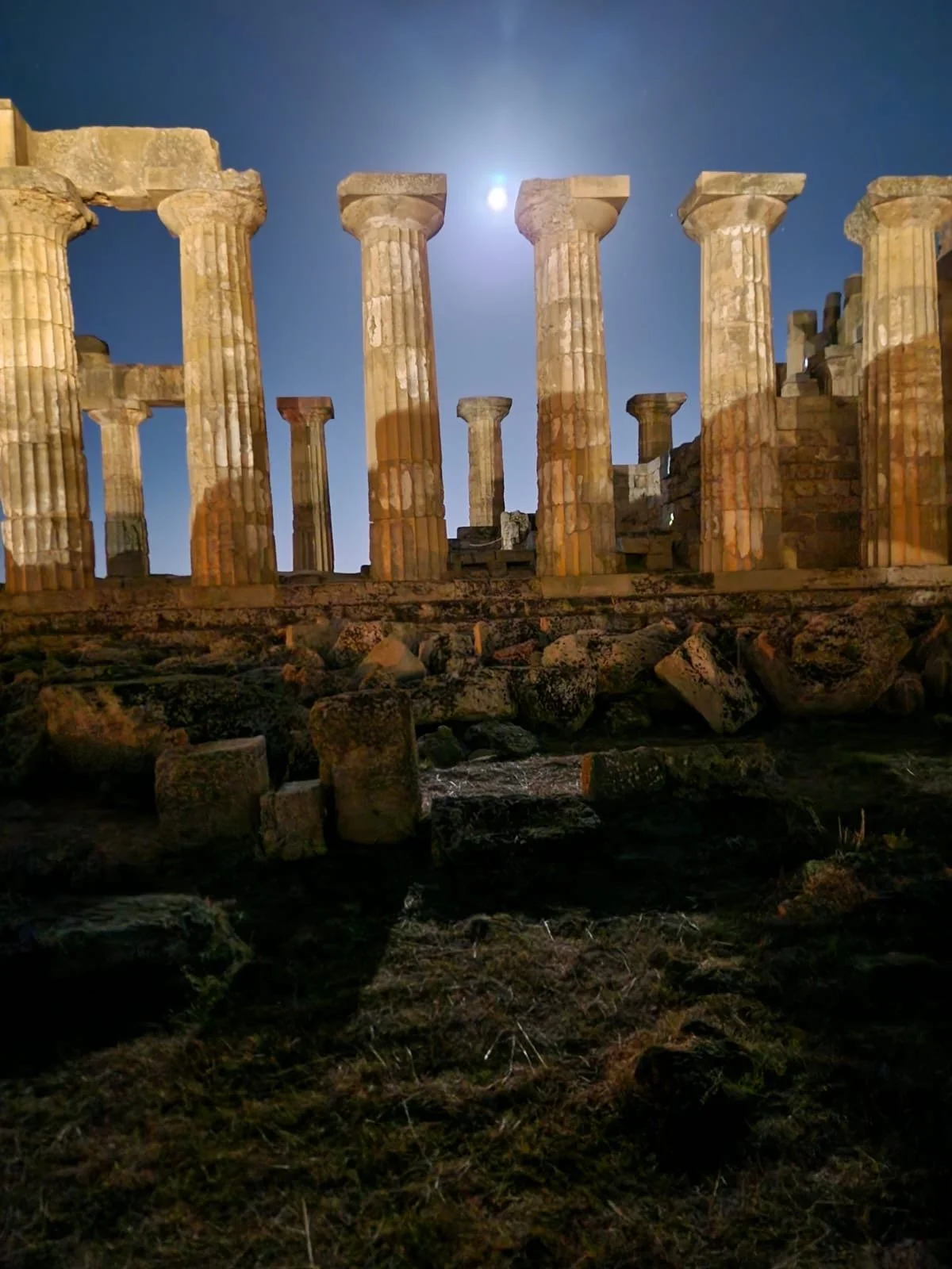 Ancient Greek ruins under a bright moon, with tall columns illuminated against a dark sky.