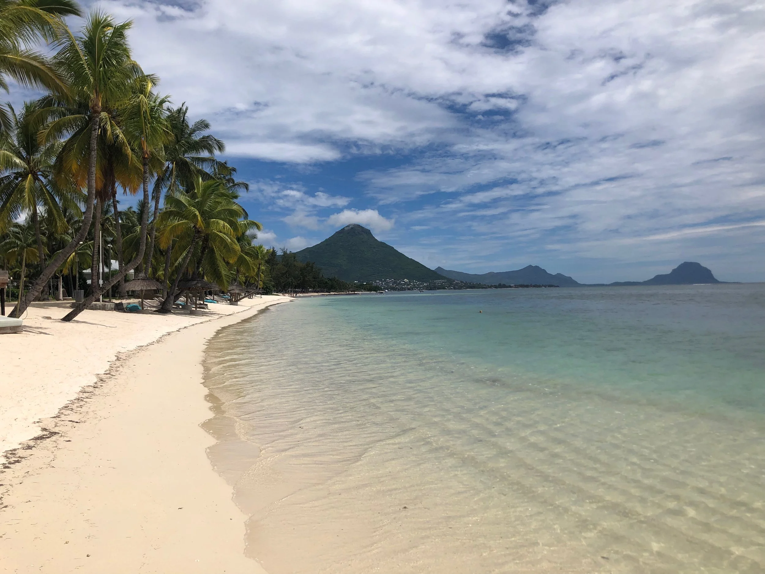 Sunny beach of Mauritius with white sand, clear water, leaning palm trees, a mountainous island in the background, and partly cloudy sky.