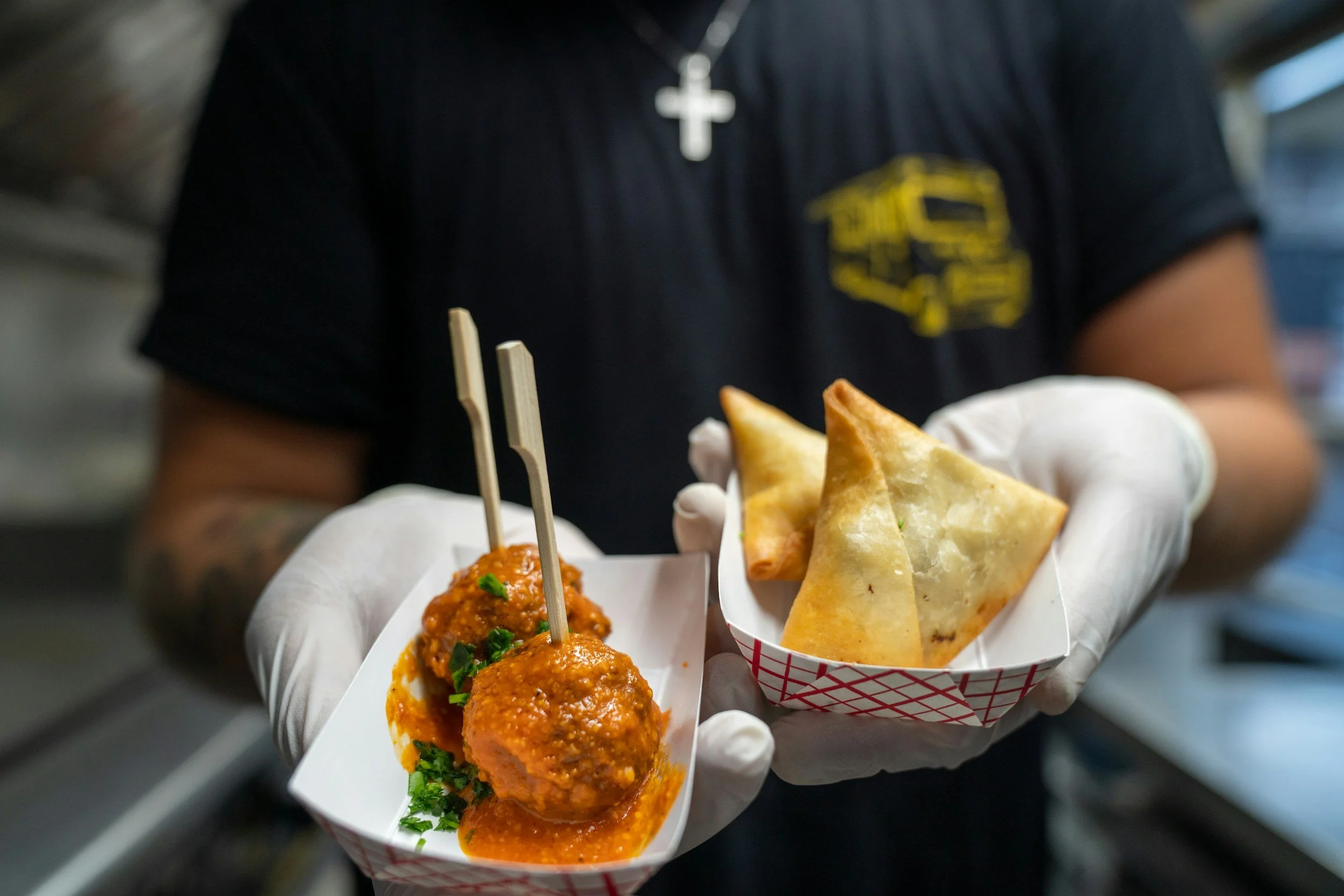 Person holding a tray with falafel balls in sauce, and a paper cup with samosas.