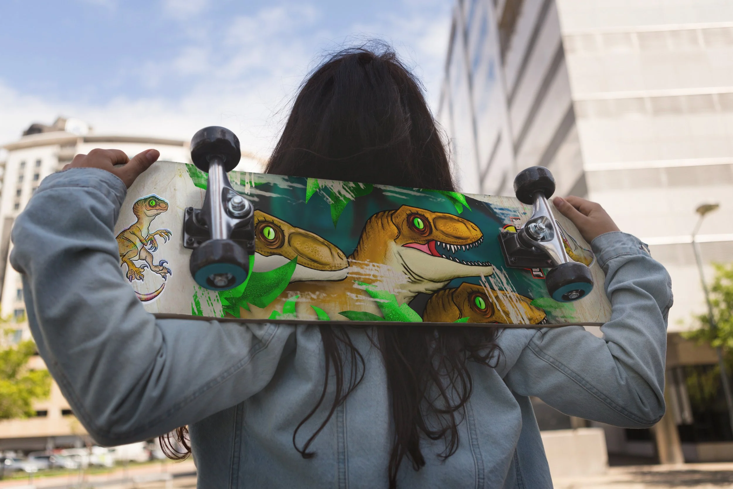 Person with long dark hair holding a skateboard with dinosaur illustrations on it, standing outdoors with buildings and blue sky in the background