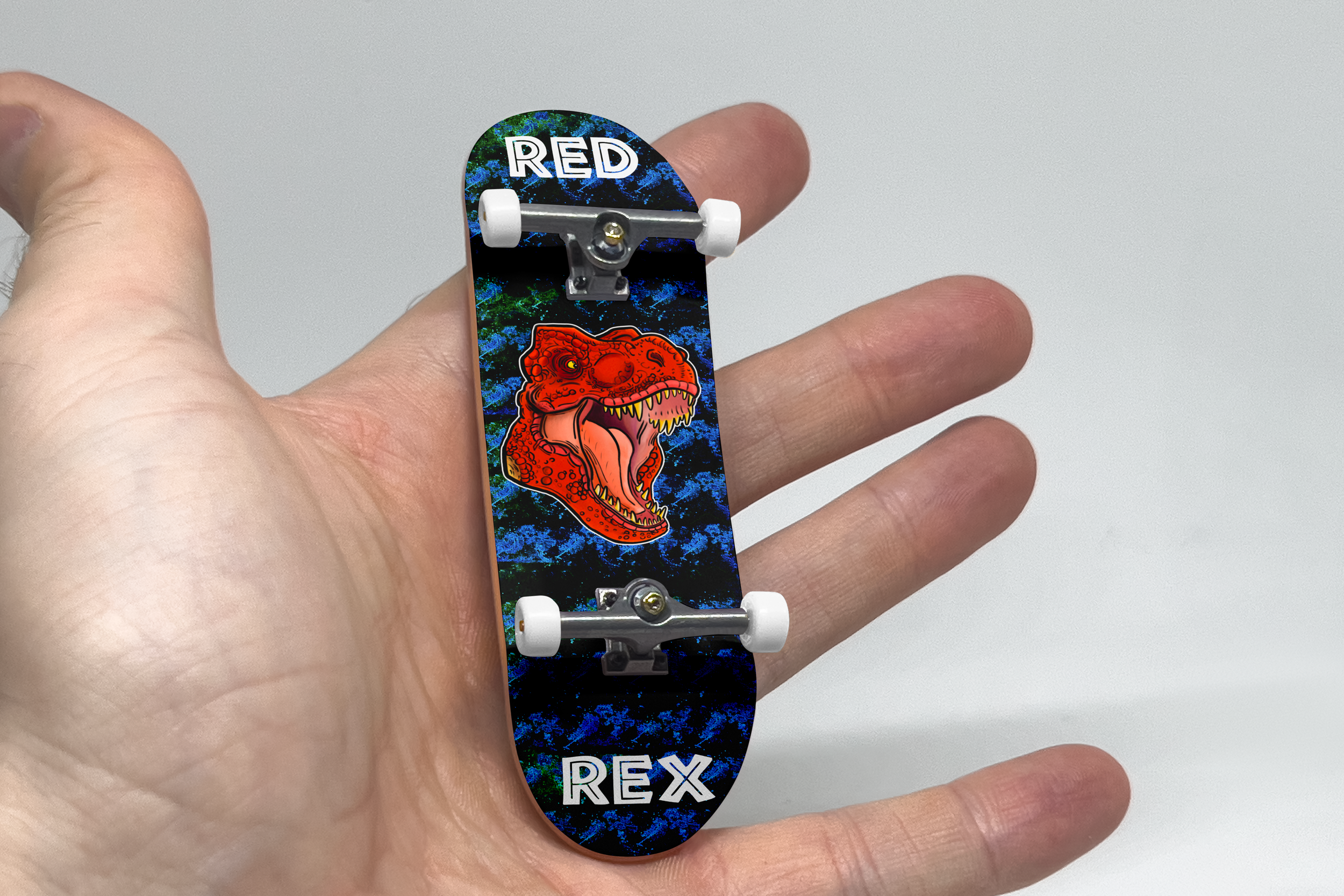Close-up of a person's hand holding a skateboard with a colorful dinosaur graphic and the word "REX" on it.