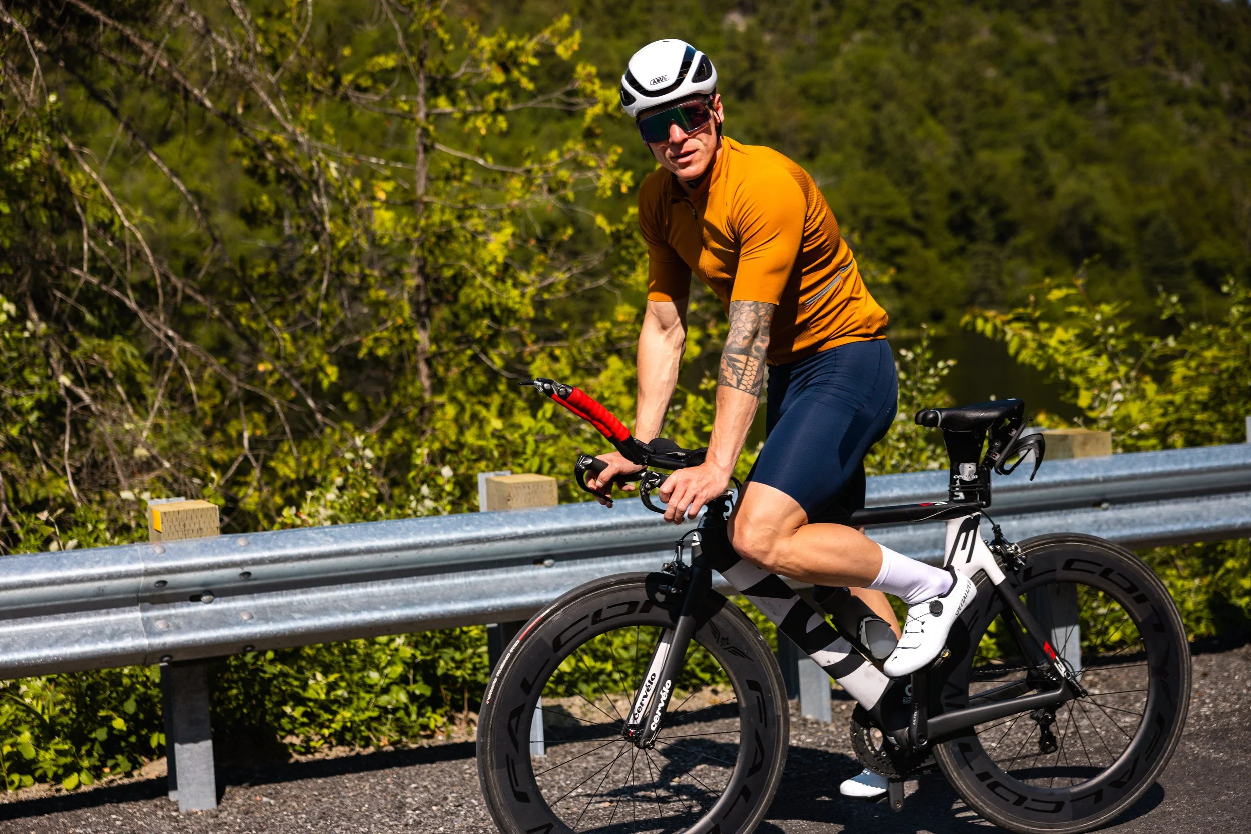 A man wearing a white helmet, dark sunglasses, an orange cycling jersey, and black cycling shorts riding a black and white road bike along a forested path with a metal guardrail.
