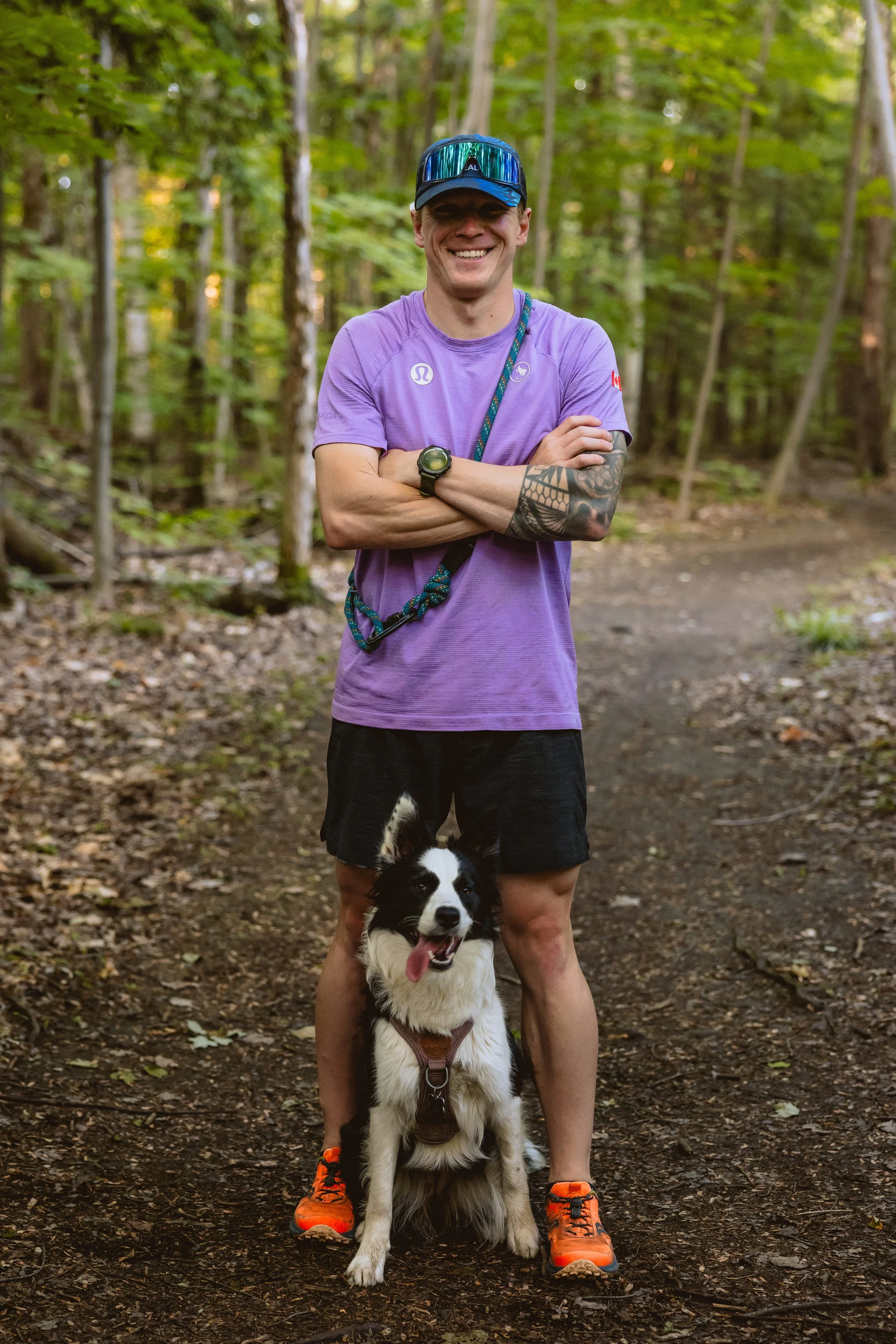A man wearing a purple t-shirt, black shorts, orange running shoes, sunglasses, and a cap stands in a forest with a black and white Border Collie dog sitting in front of him.