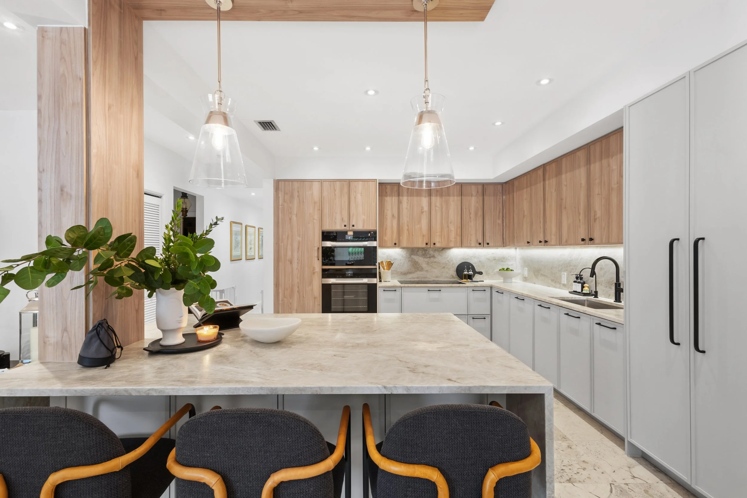 Modern kitchen with wooden cabinets, white walls, recessed lighting, and a marble countertop. Kitchen items include a plant, bowls, candles, and black soap dispensers.