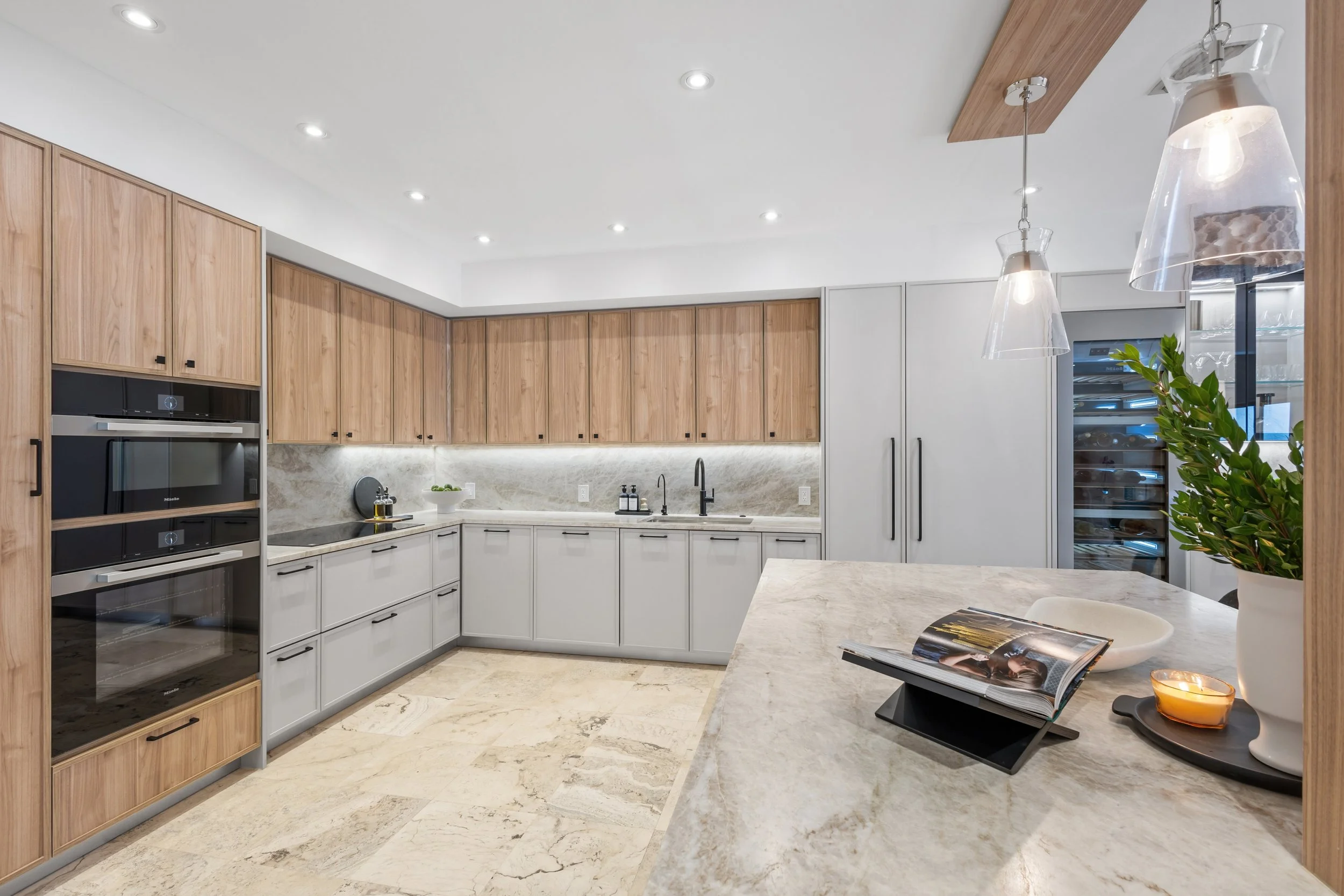 Modern kitchen with white cabinets, wooden upper cabinets, a gray marble backsplash, and a marble countertop. Recessed ceiling lights and two glass pendant lights hang over the counter. A potted plant and an open magazine are on the counter.