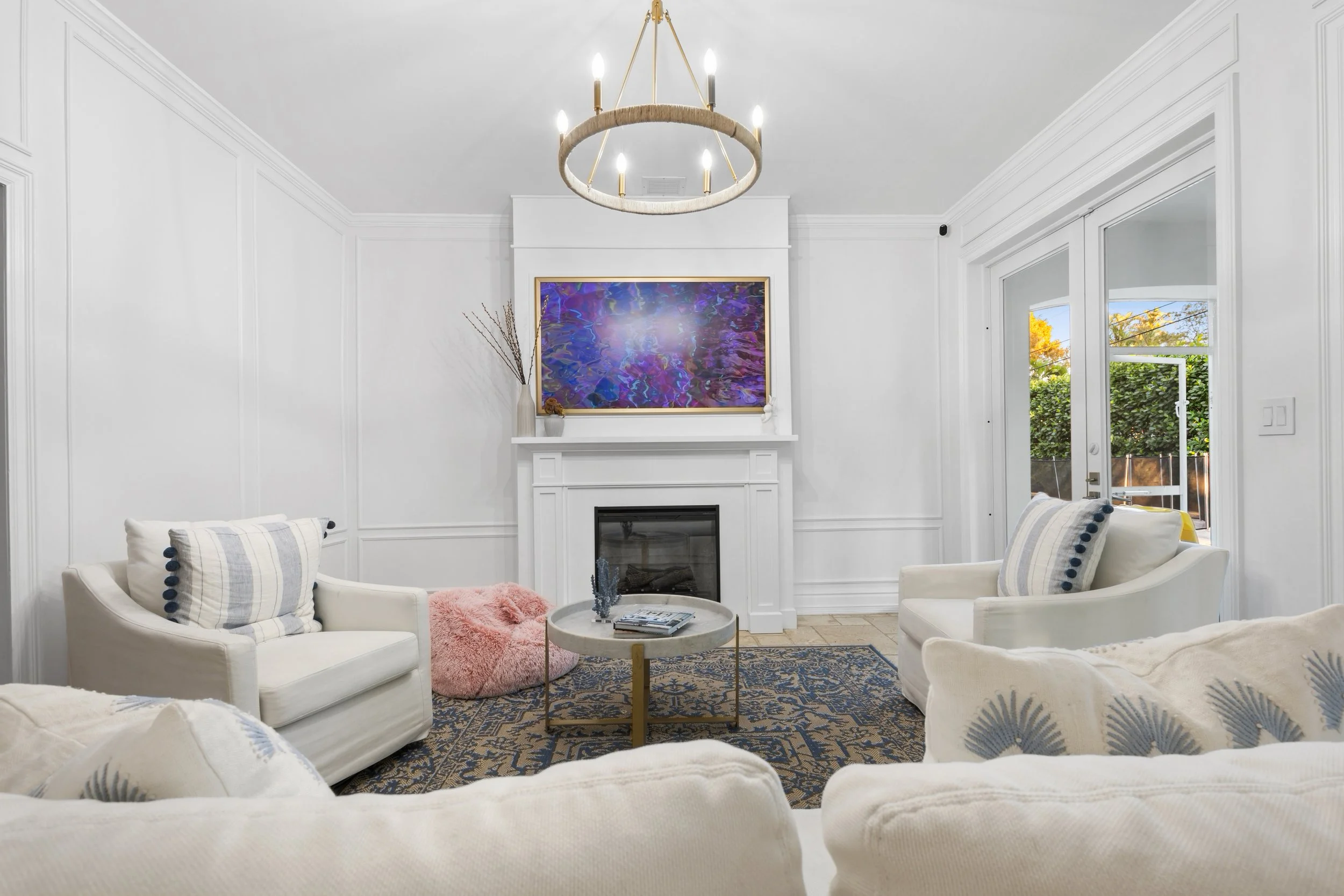 Bright living room with white walls, a fireplace, and a chandelier. Contains white couches with patterned pillows, a round coffee table, and a pink fluffy ottoman. Artwork hangs above the fireplace, and windows reveal trees outside.