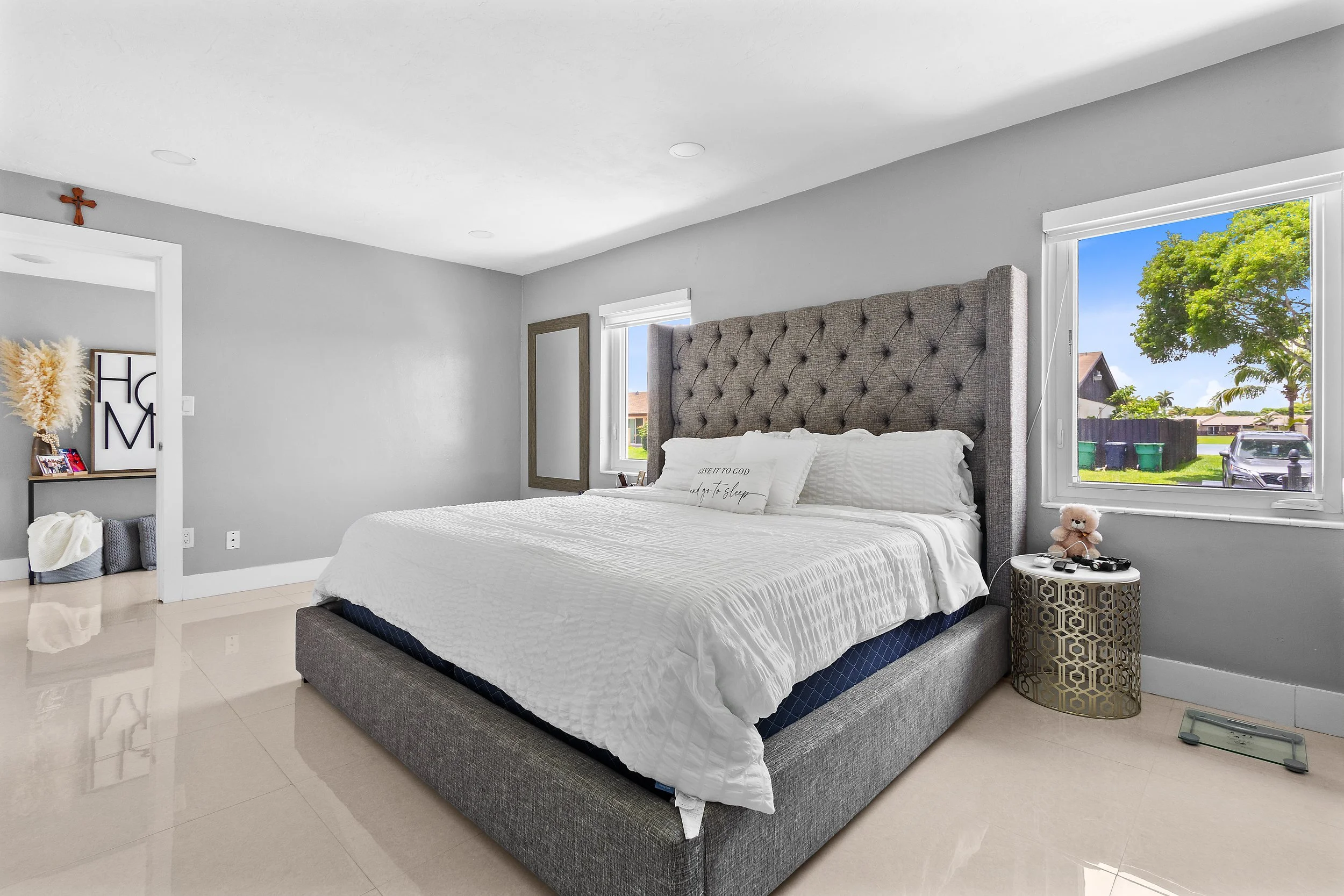 A bedroom with a large bed featuring a tufted headboard and white bedding. There are two windows with views of a tree and neighborhood outside. A small teddy bear and a round patterned side table are next to the bed. The room has light-colored walls and a shiny tile floor.