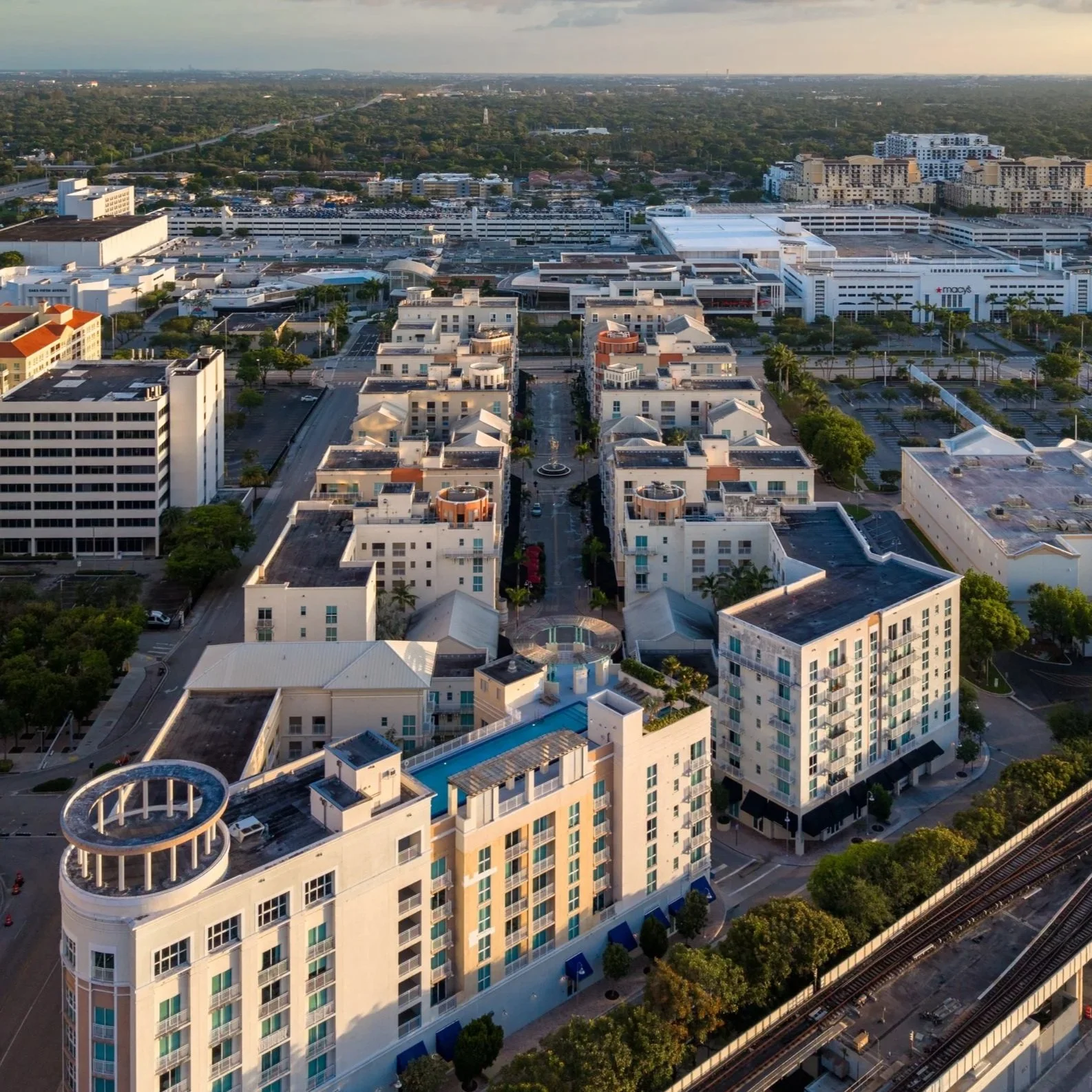 Aerial view of a cityscape featuring a large shopping mall, surrounding high-rise residential buildings, and a train track with trees lining the streets.