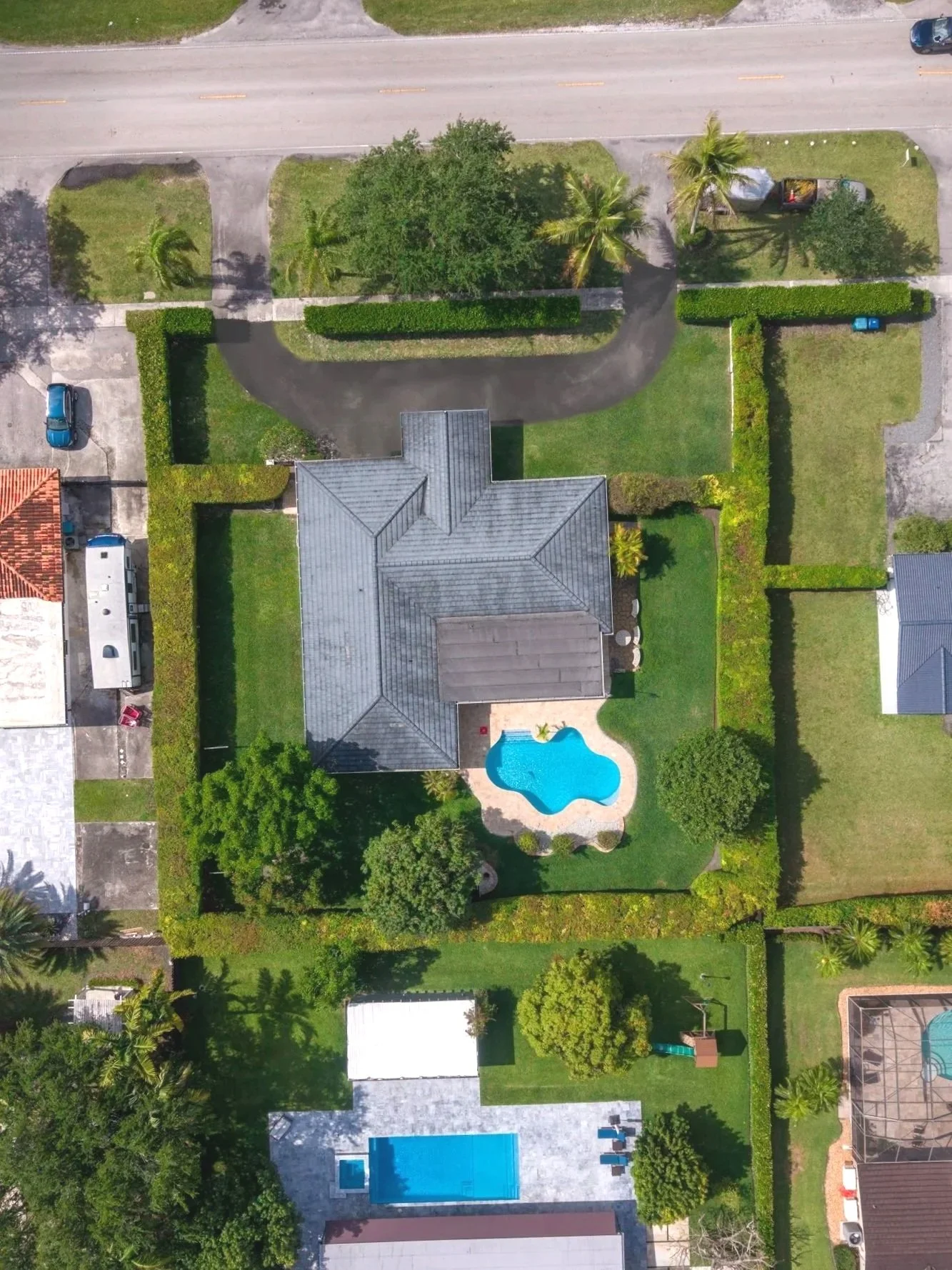 Aerial view of a residential backyard with a pool, fenced-in yard, lawn, trees, and neighboring houses.