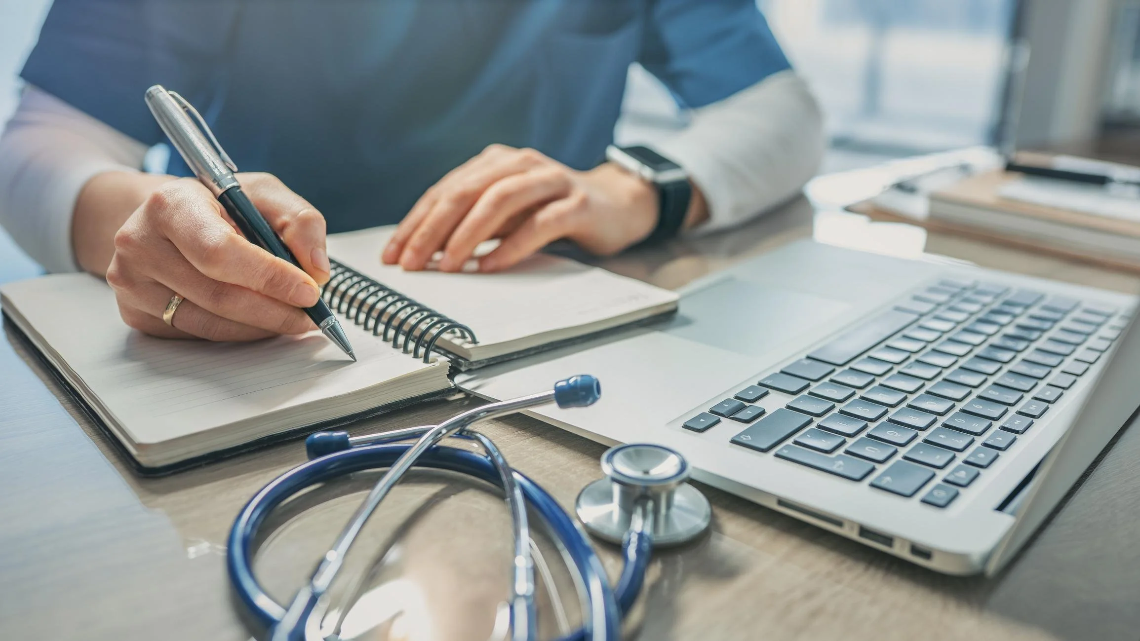 A person writing in a notebook with a pen, surrounded by a laptop, a stethoscope, and other office supplies.