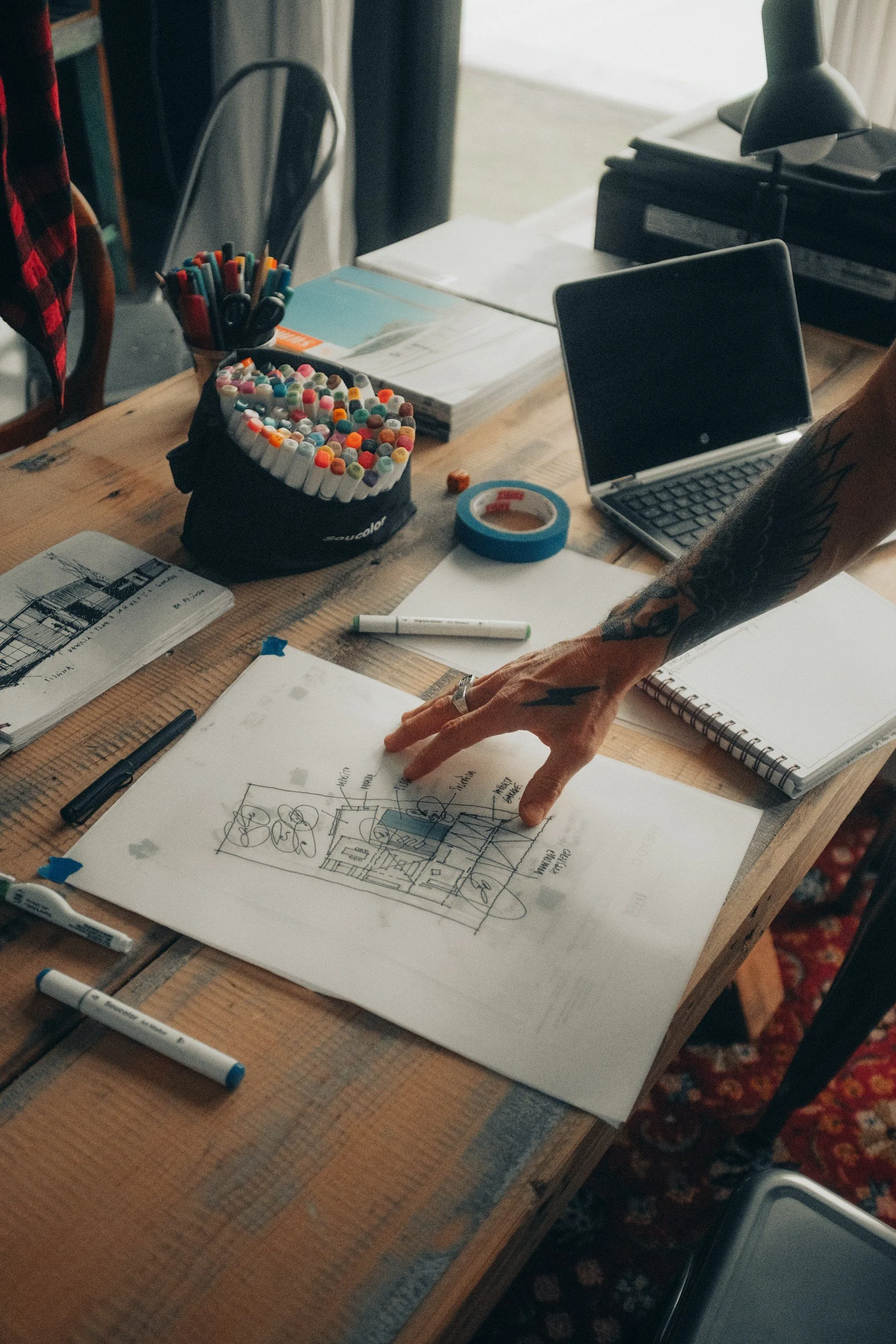 A workspace with a wooden table holding various items including a notebook with a drawing, markers, a pen, and tape. There is a laptop, a person with tattoos, and organized documents.
