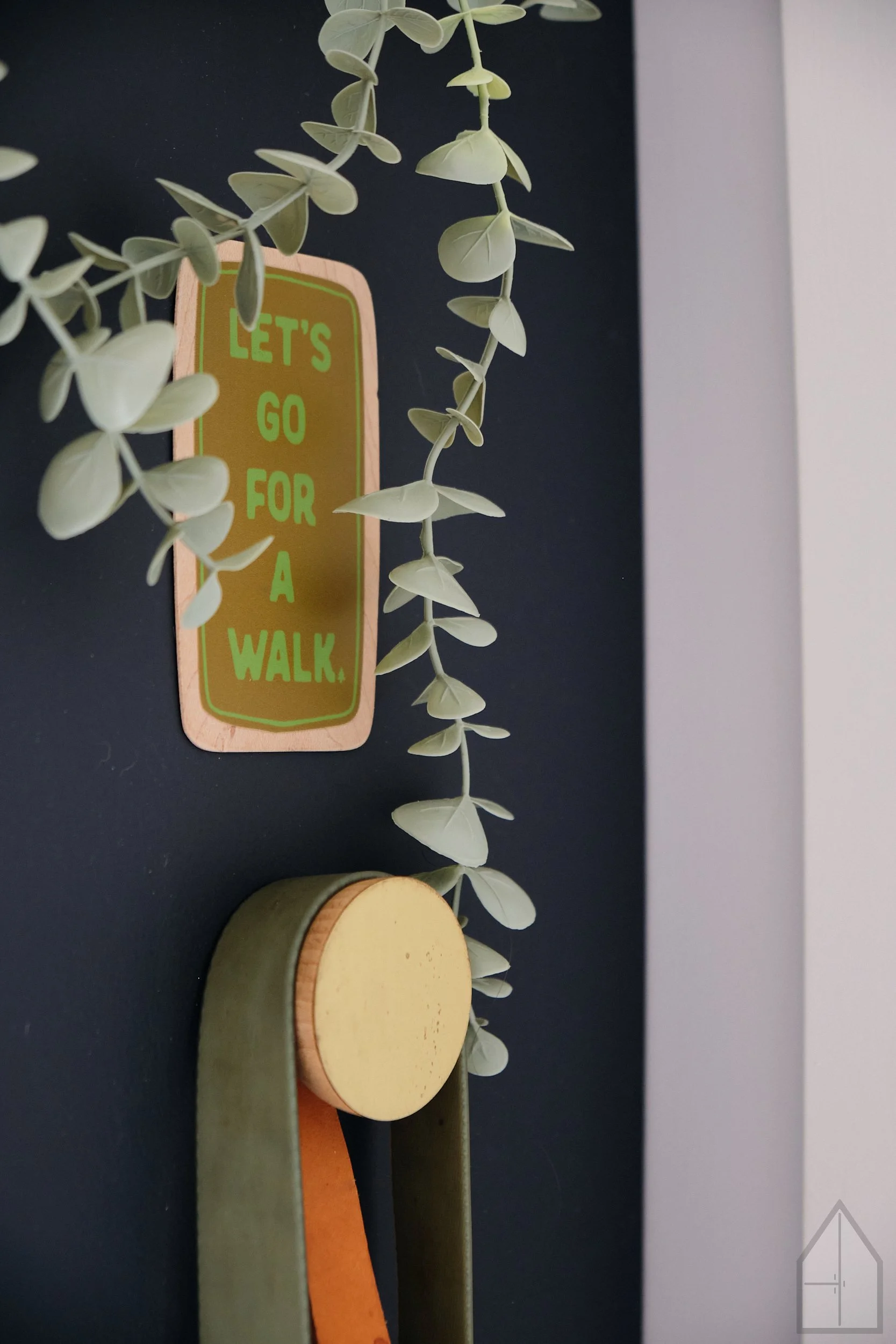 Decorative hanging plant with a sign that reads 'Let's go for a walk' attached to a black wall. The plant has trailing green leaves, and there are round wooden and orange-colored objects below.