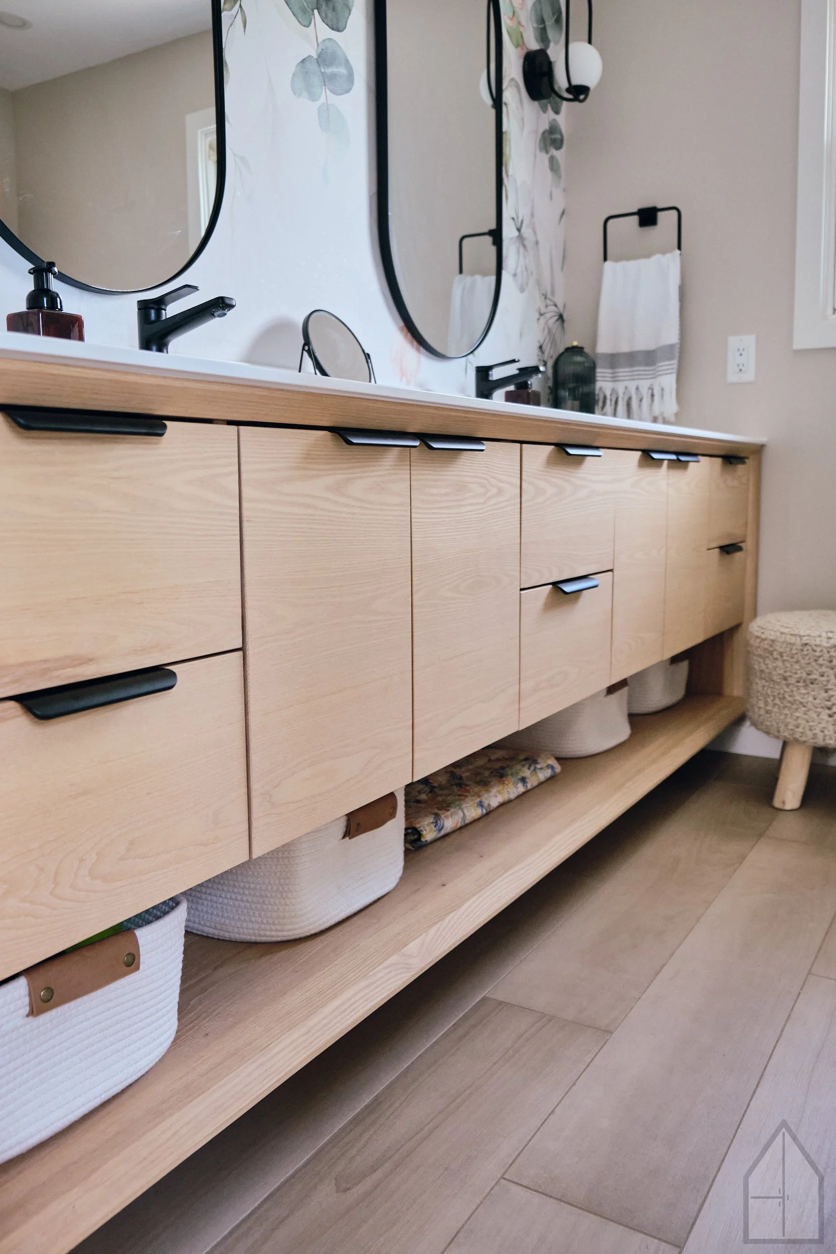 Bathroom vanity with a light wood finish, black fixtures, a large mirror, towels on a wall-mounted rack, and woven baskets and folded textiles on the lower shelf, with tan tiled flooring.