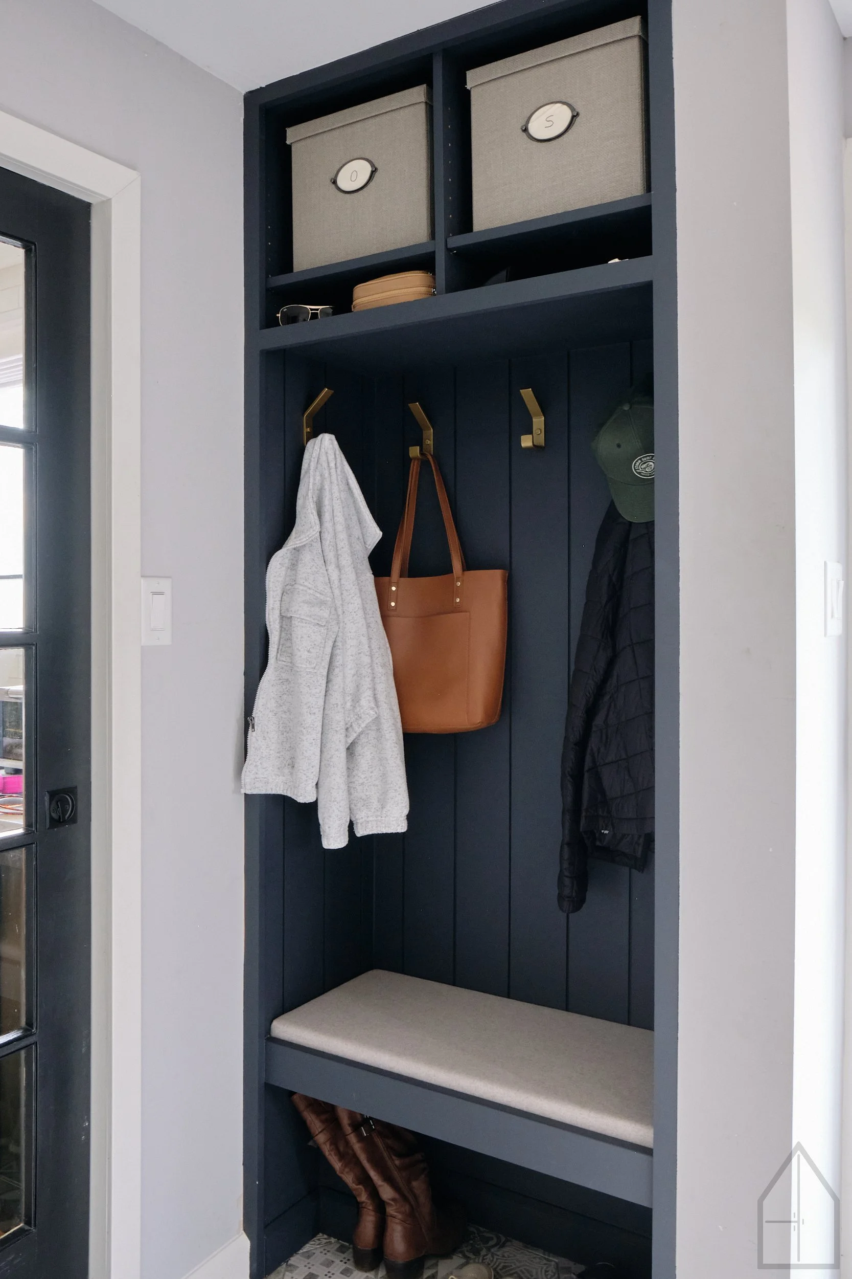 A navy blue wall-mounted entryway organizer with four hooks, holding a gray hoodie, a tan leather handbag, and a green cap. Two cubby spaces at the top contain gray storage bins labeled with the numbers 0 and 5. There is a pair of brown boots on the floor below the organizer.
