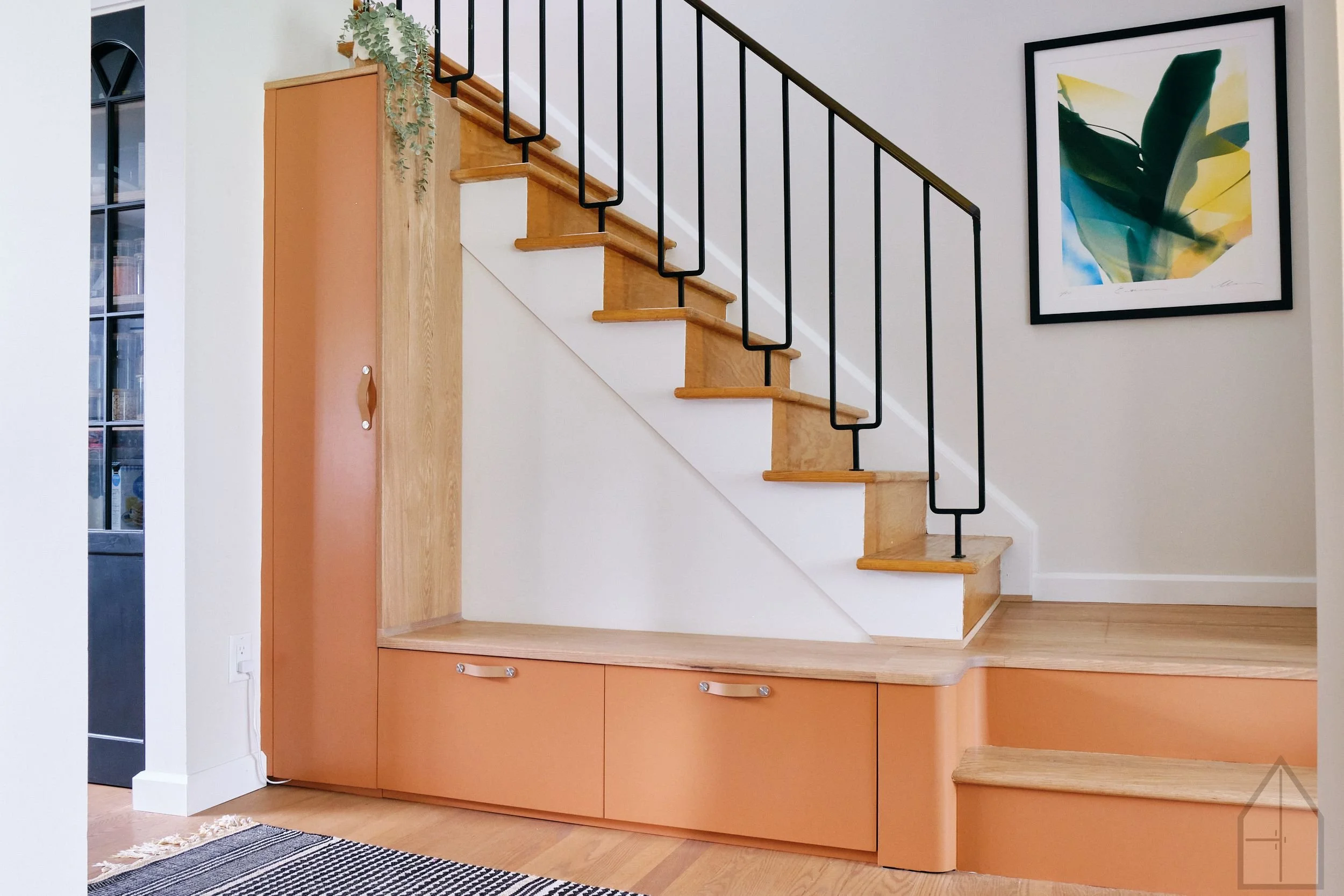 Interior of a modern home staircase with wooden steps, black metal railing, a framed abstract art print on the white wall, a tall cabinet with a folding door, and a narrow window on the left side.