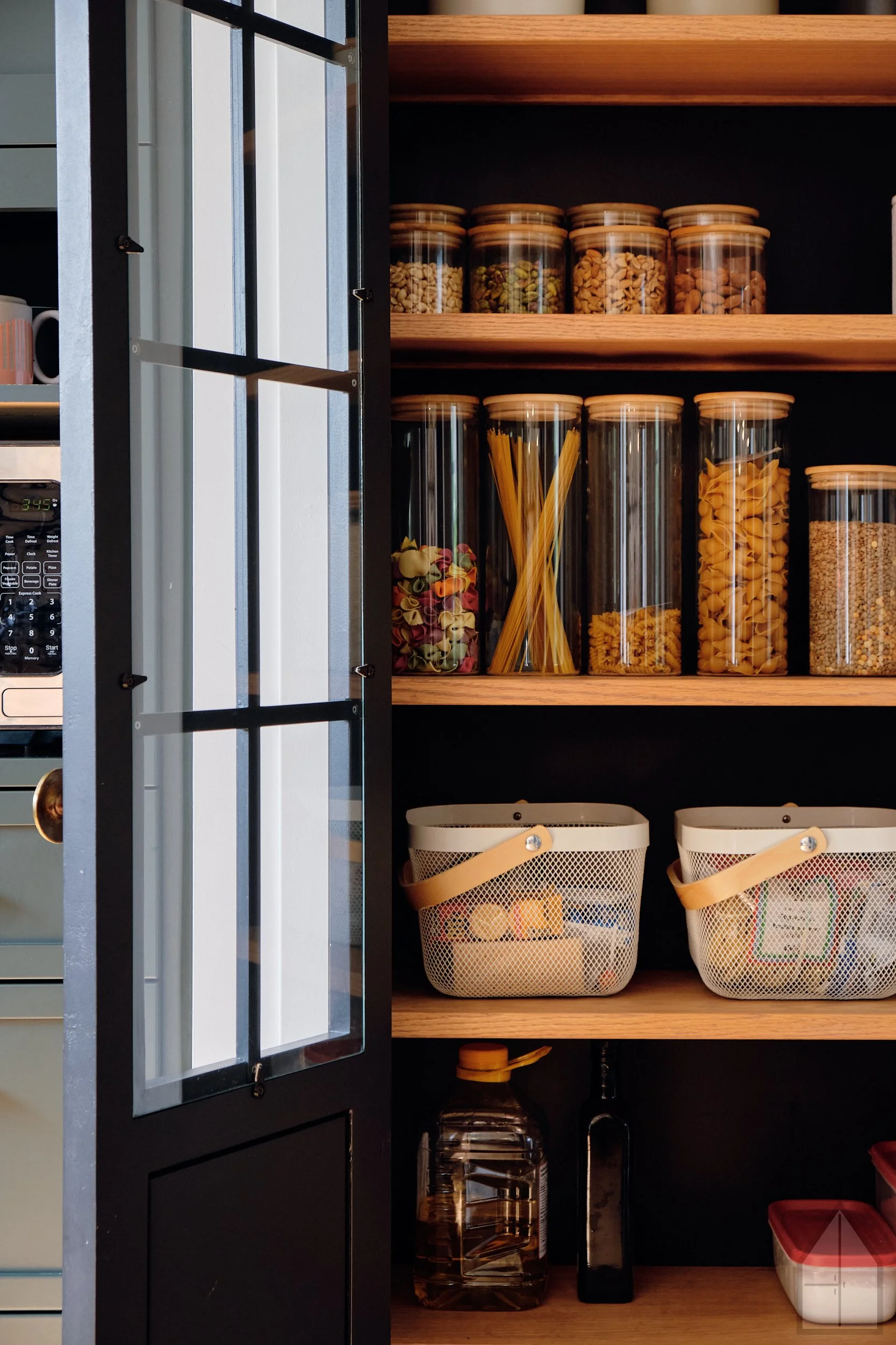 Open black kitchen cabinet with glass doors, containing jars of pasta, nuts, and snacks, and white baskets on the lower shelf, with a microwave partially visible on the side.
