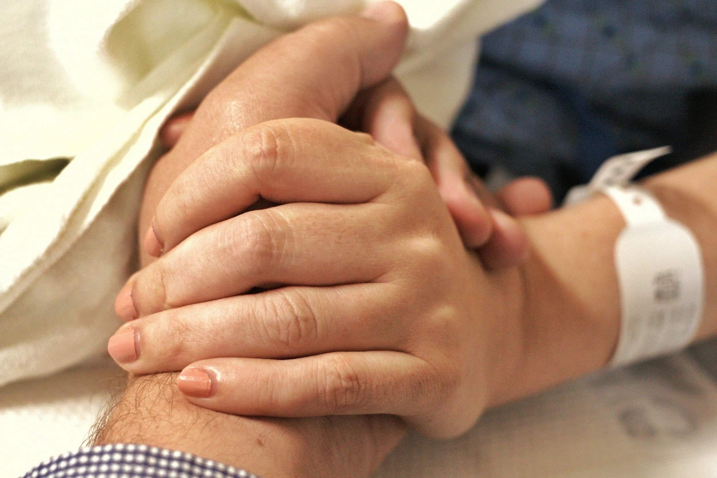 Close-up of two hands clasped, one appearing to be a patient's hand with a hospital identification bracelet. The background includes part of a person's arm with a checkered sleeve.