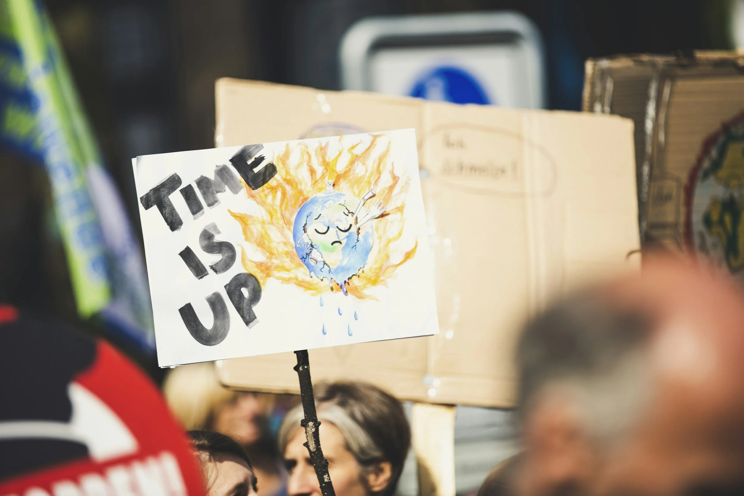 A protestor holding a sign with a drawing of the Earth on fire with tears, a thermometer in its mouth, and the words 'Time Is Up' in bold black letters.