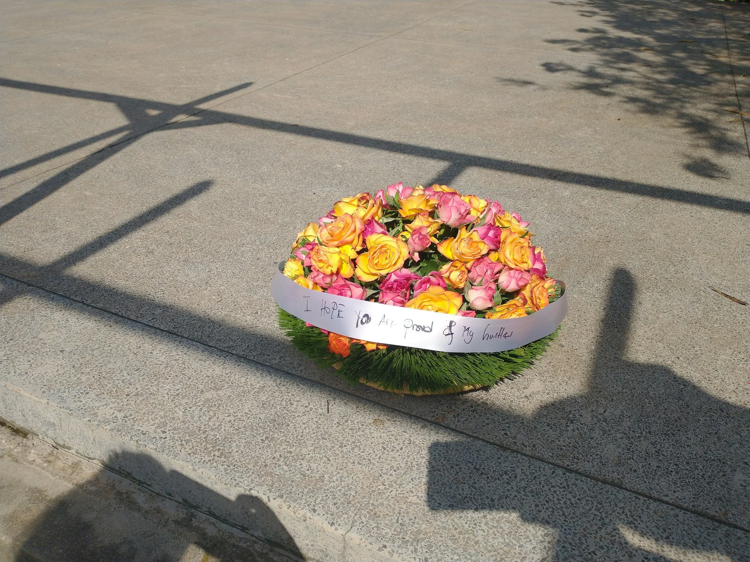 A bouquet of pink and yellow roses with a white ribbon that has handwritten text on it, placed on a concrete surface with shadows of trees and a lamp post.