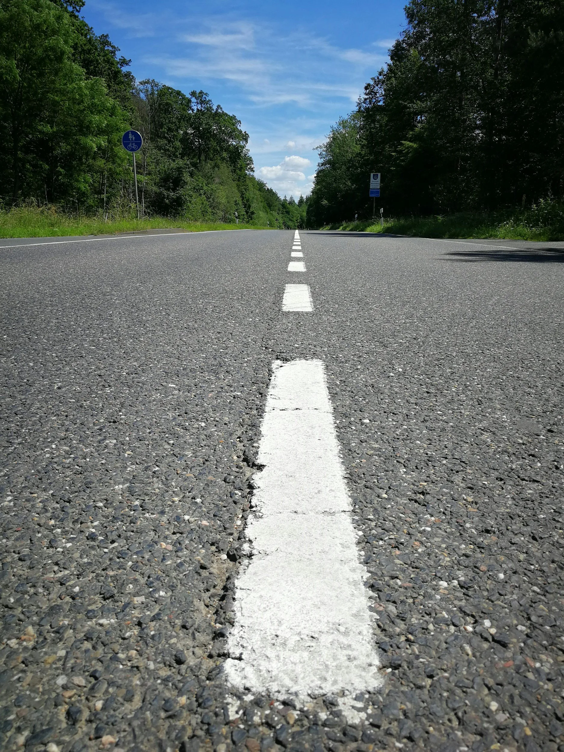 A straight asphalt road with a broken white line in the middle, surrounded by green trees under a blue sky with some clouds.