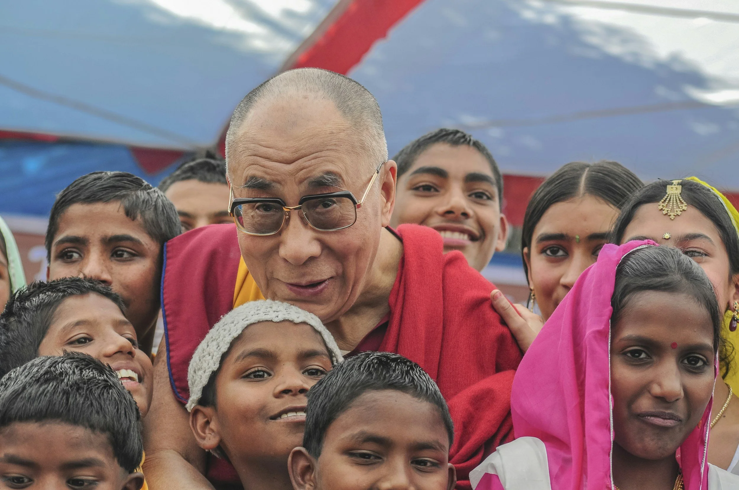 A group of children and an older man, who is likely a monk or spiritual leader, gathered closely together outdoors, with a colorful canopy or banner overhead.