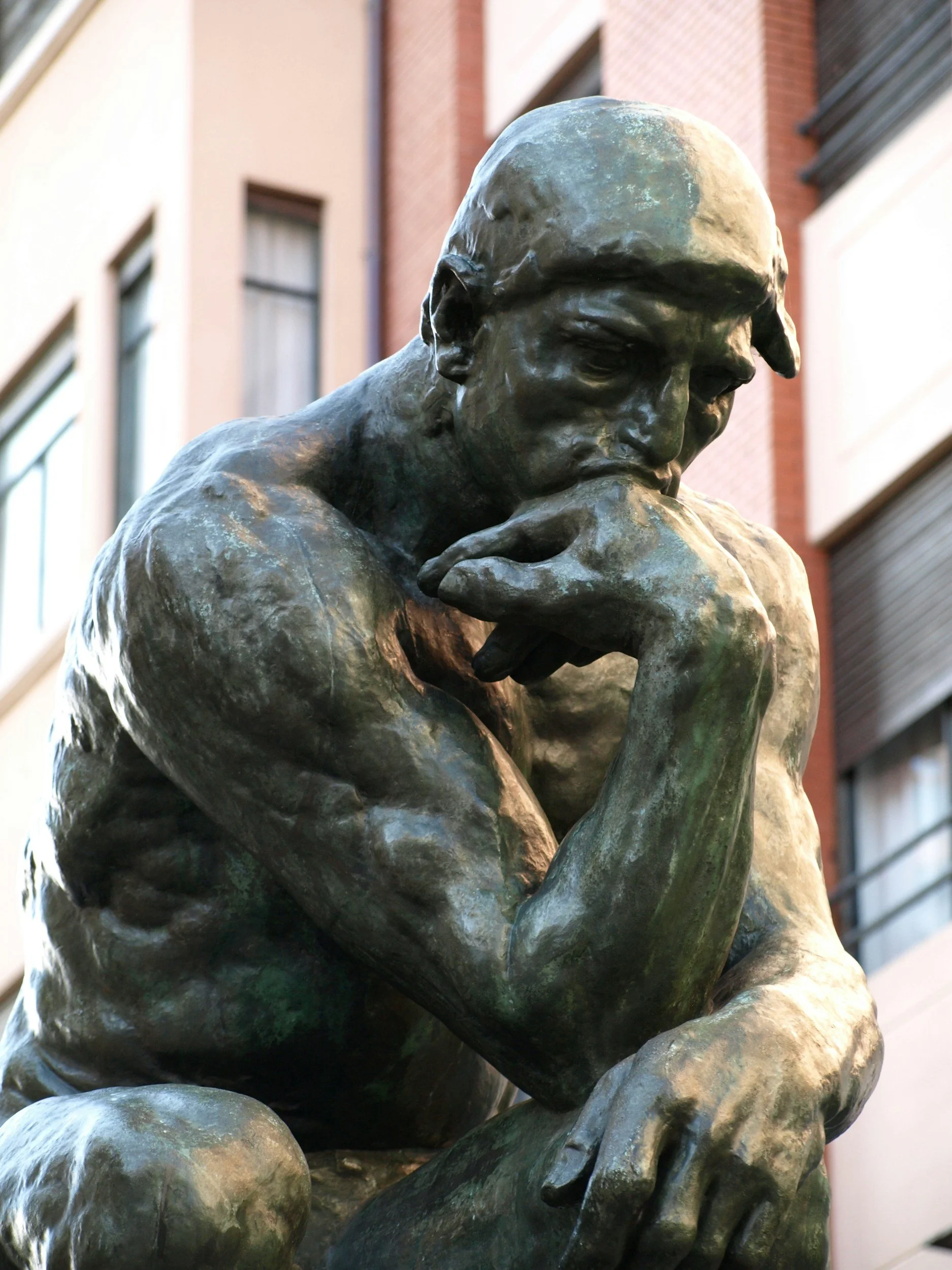 Close-up of a bronze statue of a contemplative man with his hand on his chin, set against urban buildings.