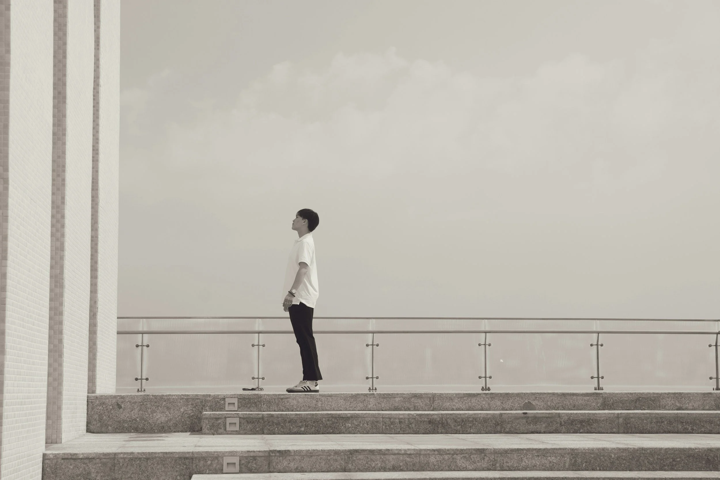 A young man in a white shirt and black pants stands on steps, looking upward against a gray sky with soft clouds.