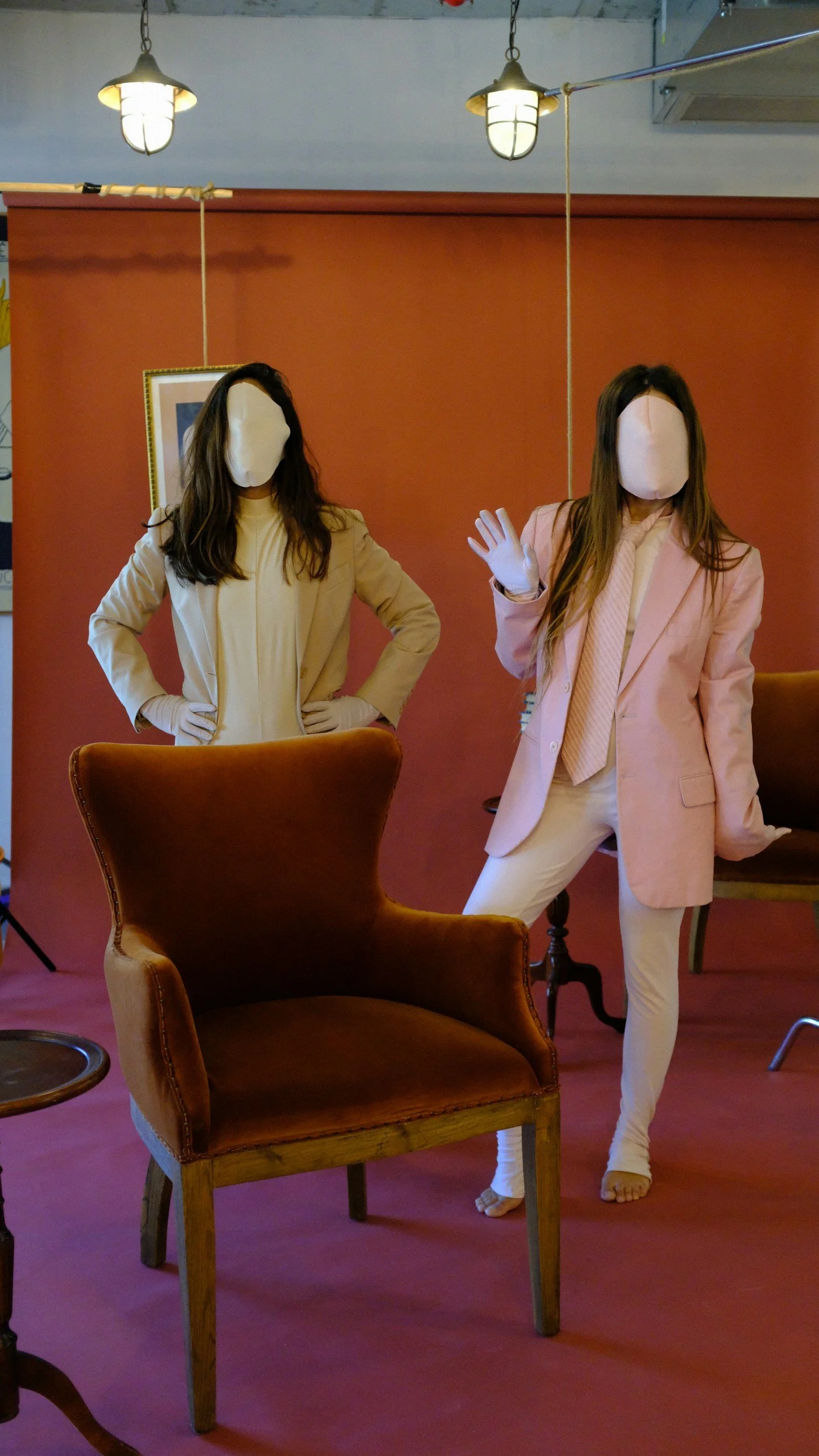 Two mannequins with faceless heads dressed in beige and pink suits, standing in a room with warm lighting and existing furniture, including a brown armchair and a wooden chair, with a red backdrop and hanging lights.