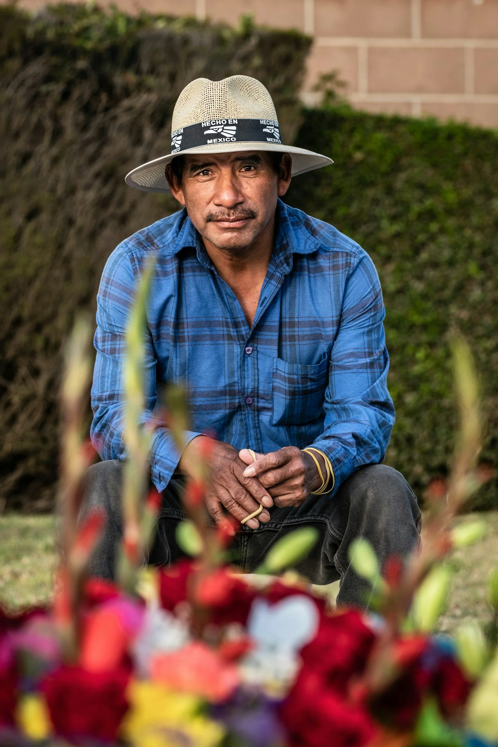 A man wearing a straw hat with a band that says "Hecho en Mexico" and a blue plaid shirt, sitting outdoors with a blurred background, surrounded by colorful flowers in the foreground.