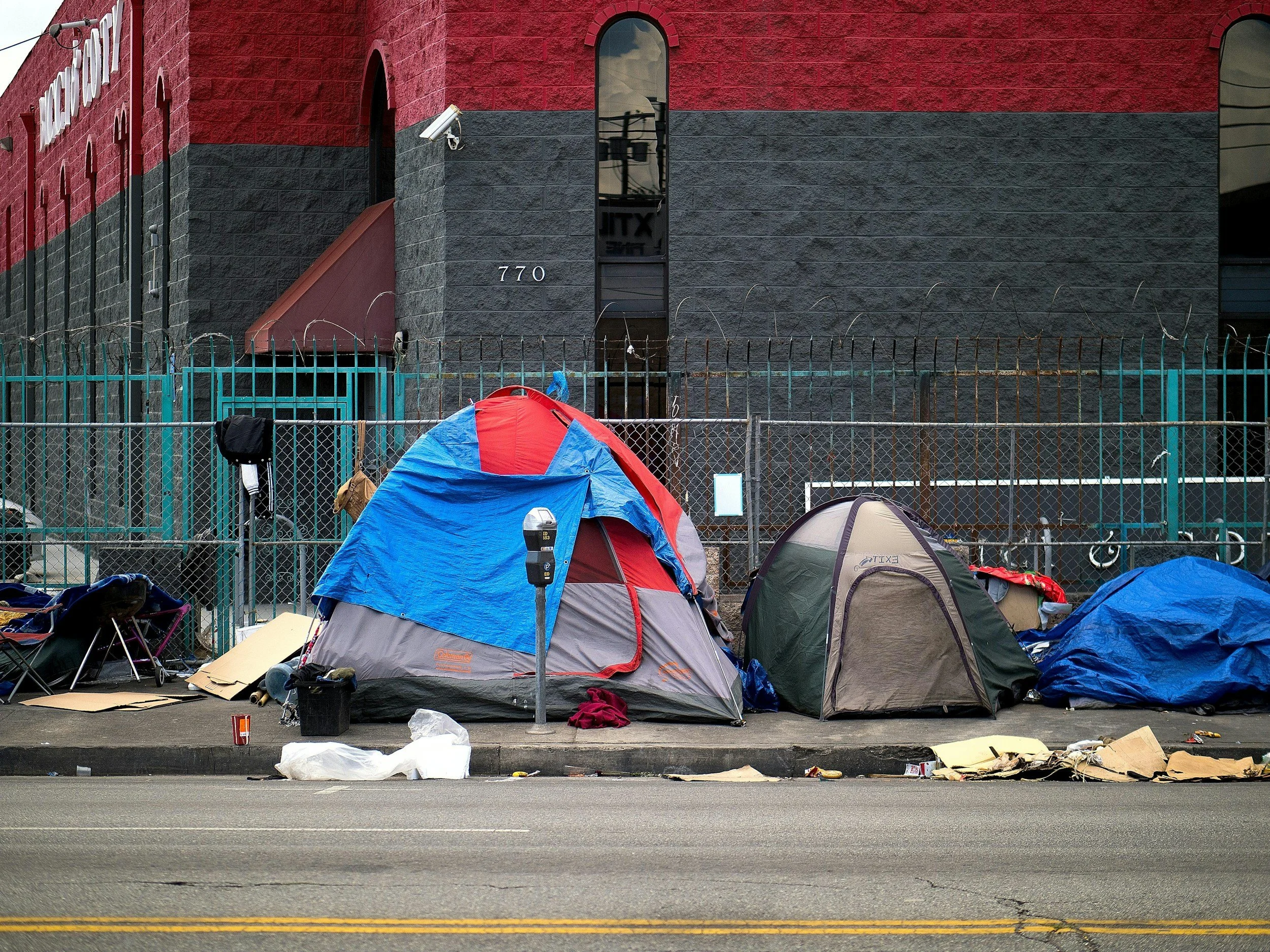 A row of tents set up along a sidewalk in front of a red and black brick building, with various personal items and trash scattered on the ground.