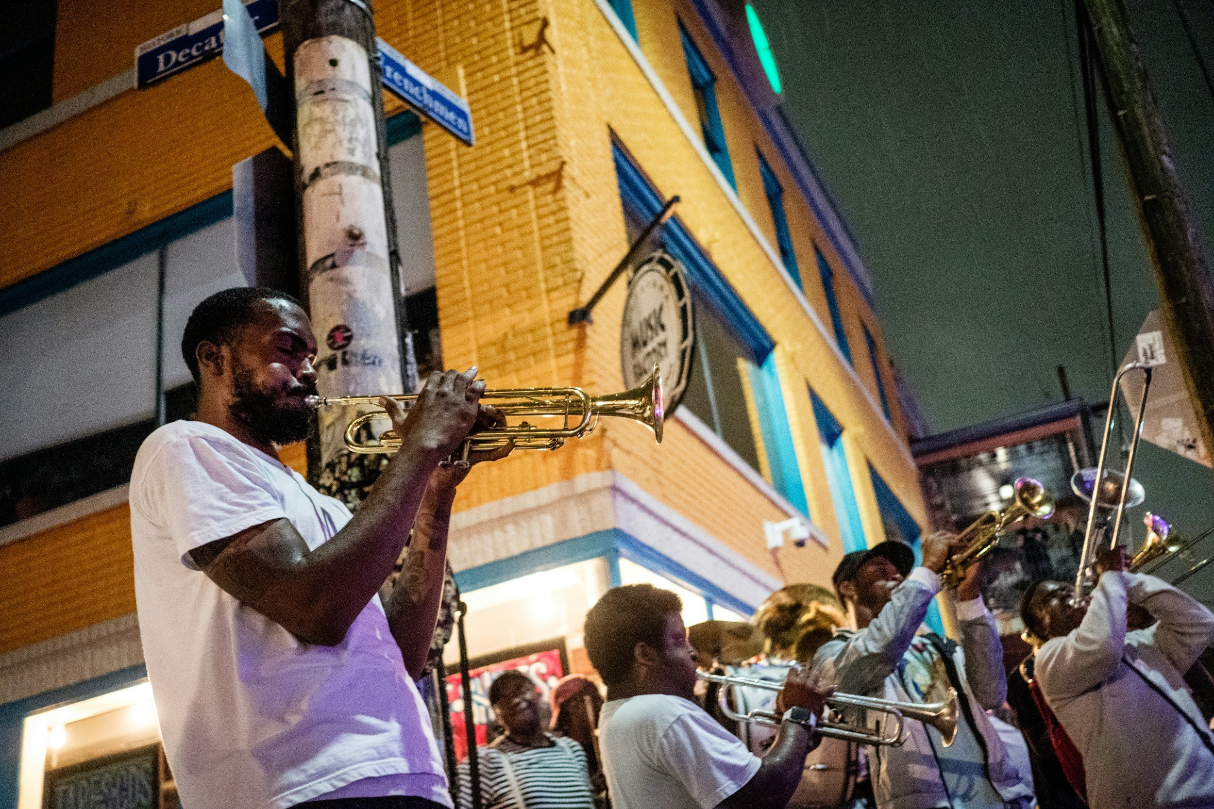 A man playing the trumpet in a group performance at night on a city street, with colorful buildings and street signs visible in the background.
