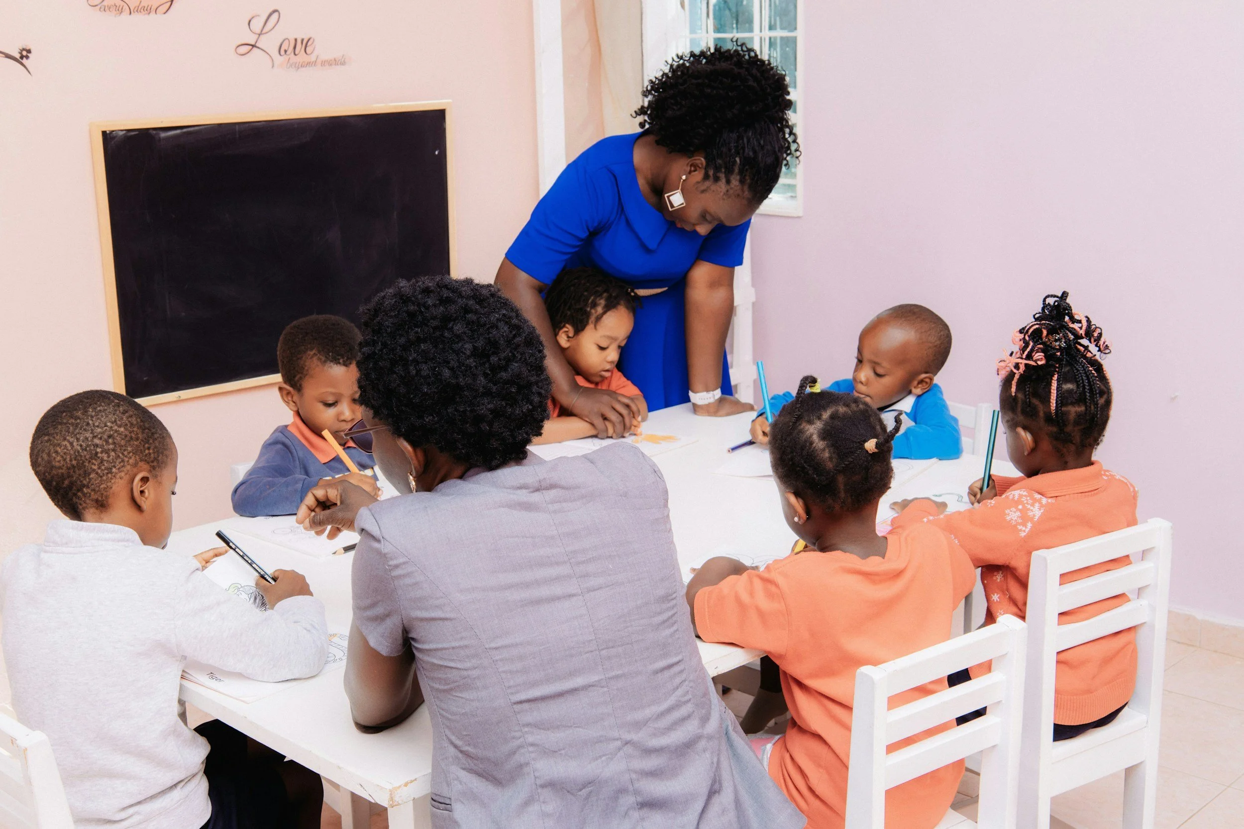 A classroom with a group of children seated at a white table, drawing on paper with colored pencils, supervised by a woman in a blue dress and large earrings, near a pink wall with blackboard and wall decals.