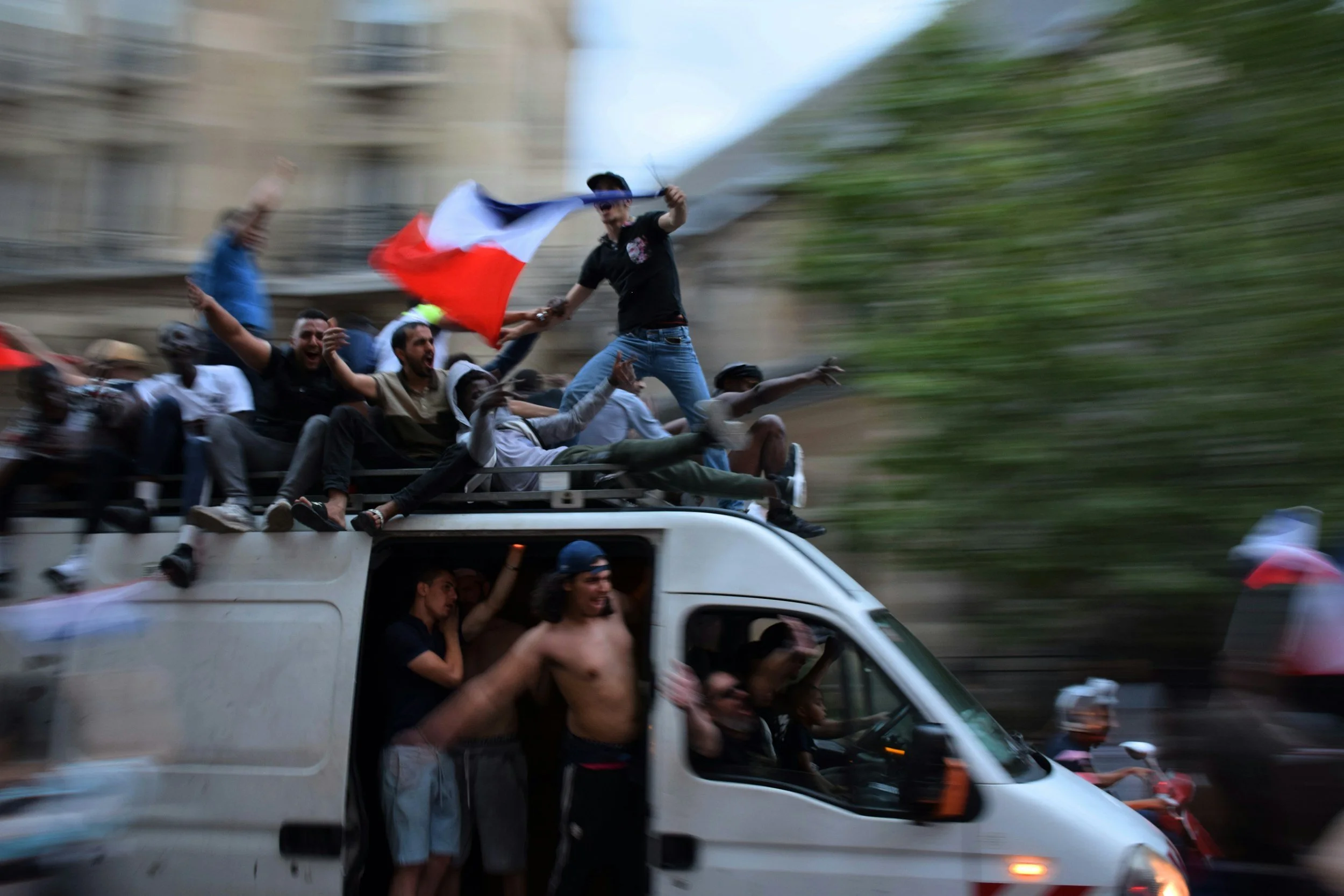 People riding on top of a white van, including a person waving a French flag, during a parade or celebration in an urban area.