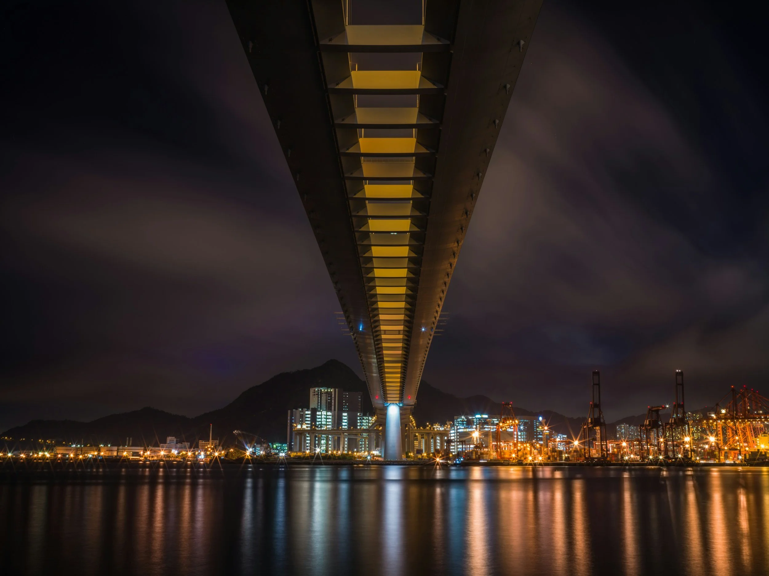 Nighttime view of a tall bridge with yellow lights reflecting on the water below, city buildings and cranes in the background, and mountains on the horizon.