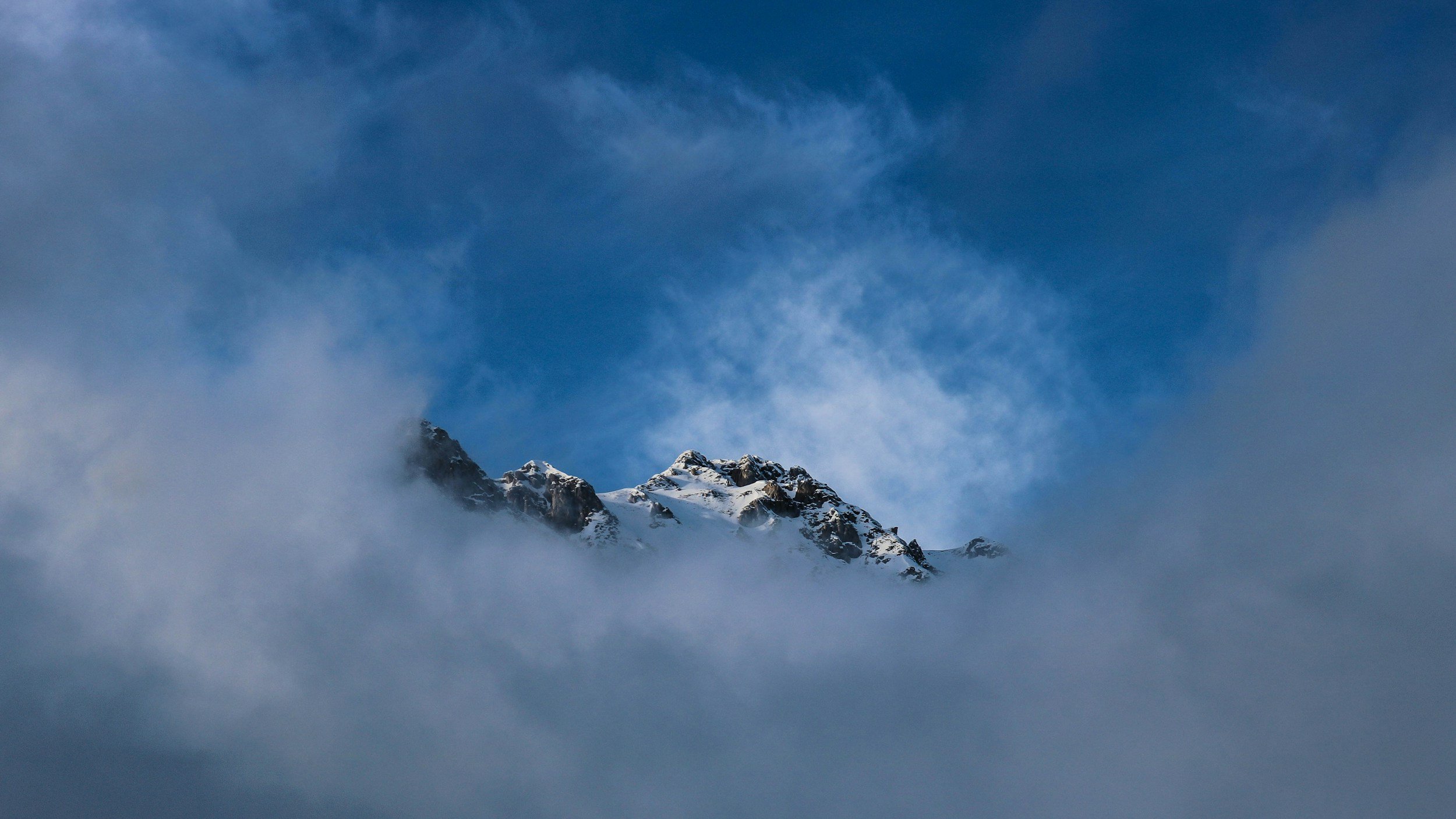 Snow-covered mountain peaks emerging through clouds against a blue sky.