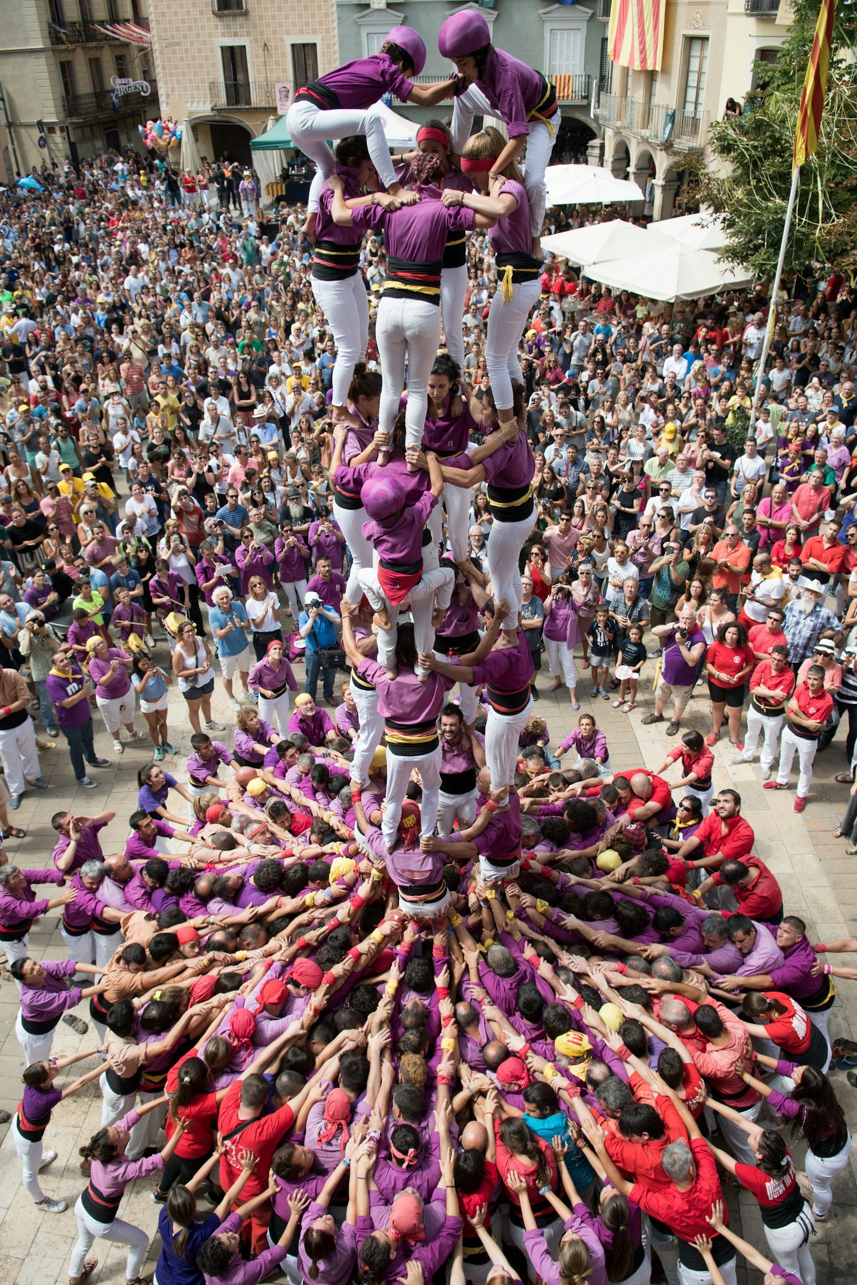 Colorful human tower, known as castells, being performed outdoors during a festival, surrounded by a crowd of spectators.