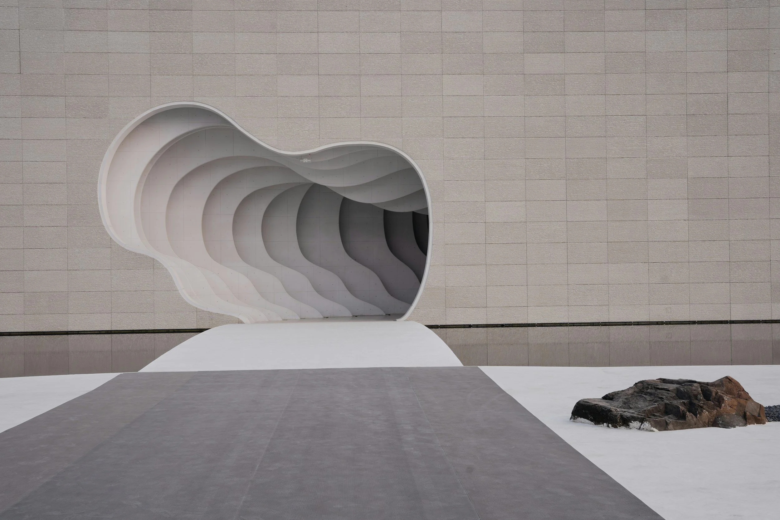 A modern architectural building with a large, irregularly shaped opening that looks like a stylized eye or abstract form. The building features curved, flowing lines and a textured beige exterior with tiled panels. There are rocks and a small landscaped area visible at the bottom right corner.