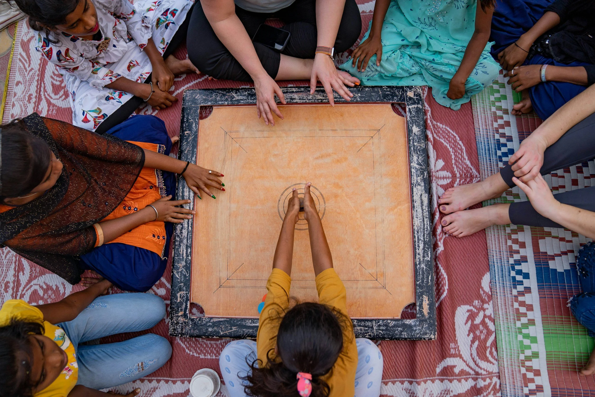 A group of children and an adult sitting cross-legged on a carpet around a rectangular game board, with the adult's hands pointing at the board.