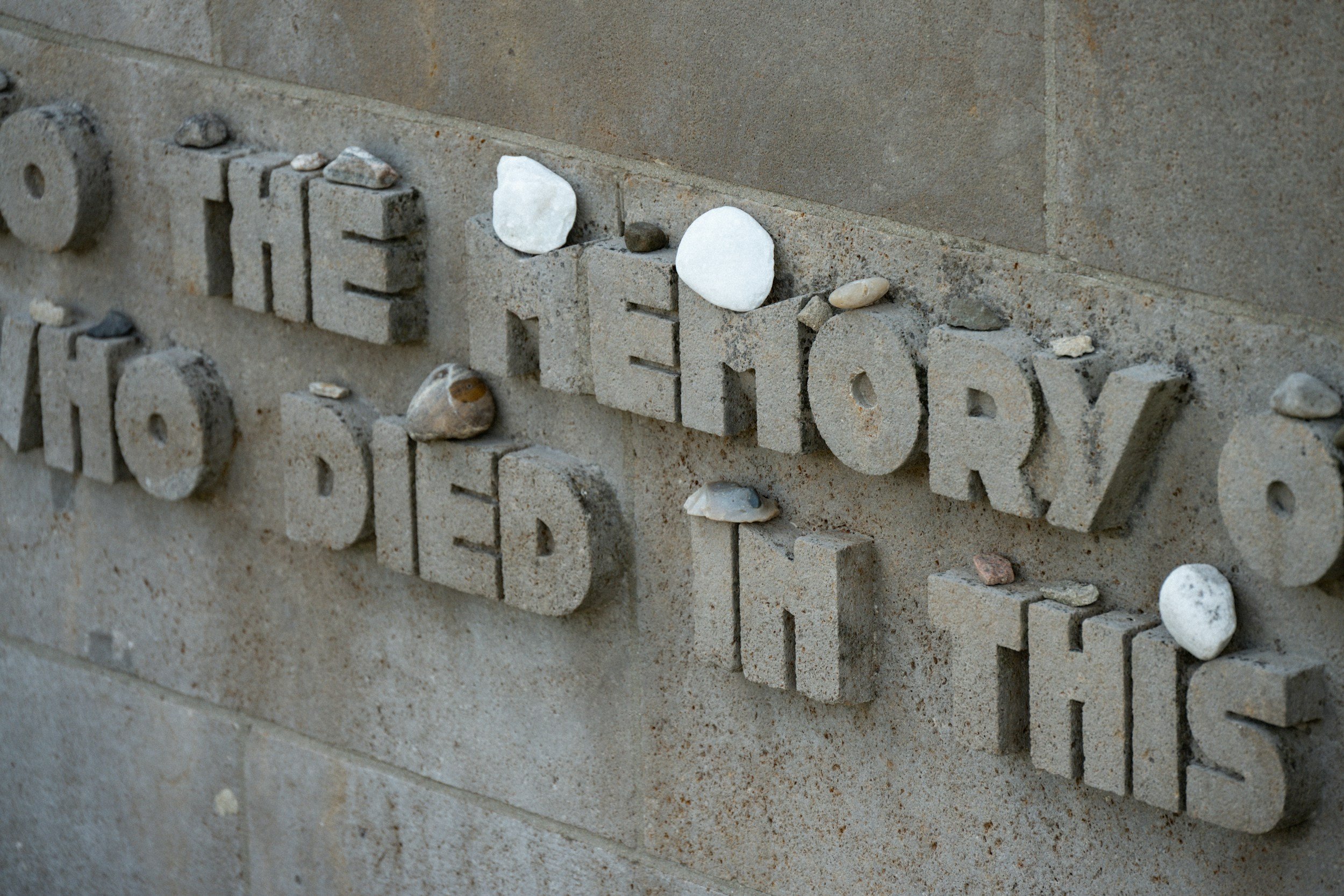 Close-up of a stone wall with letter carvings and small rocks placed on top of the carved letters.