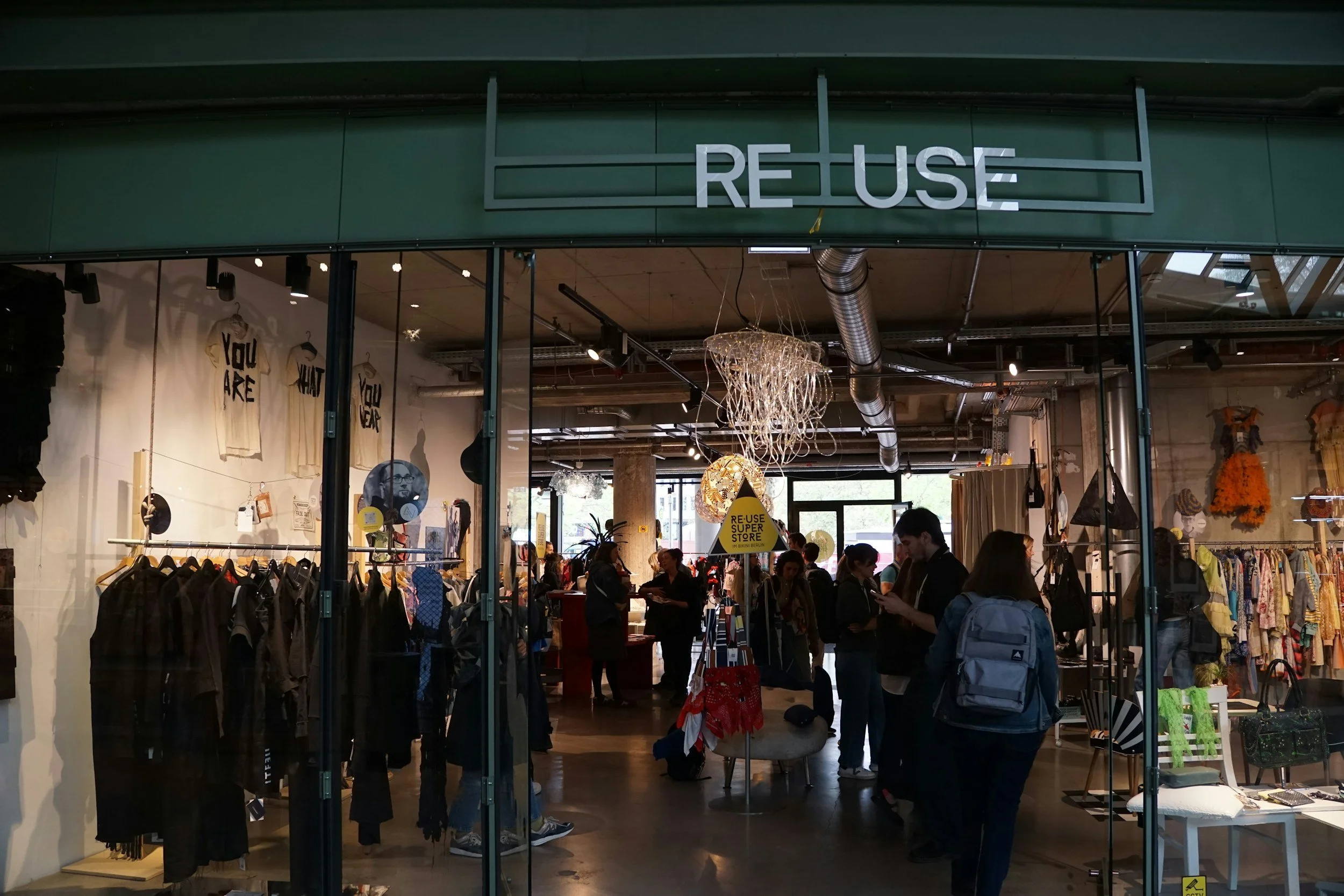 Storefront with a large sign reading 'REUSE' above the entrance, clothing and accessories inside, and several people browsing.